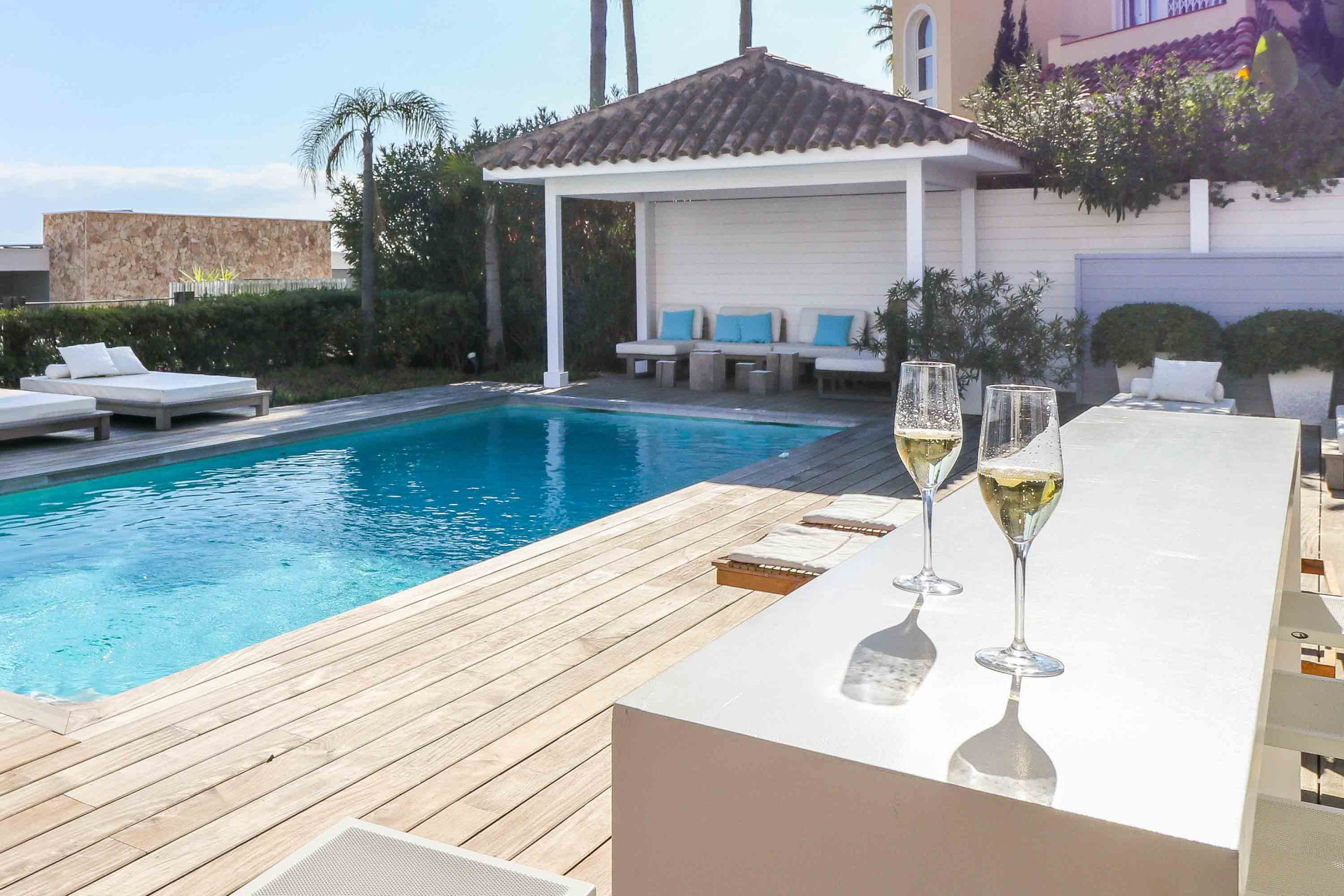 Sparkling wine glasses on a white bar counter by a swimming pool and covered lounge area.
