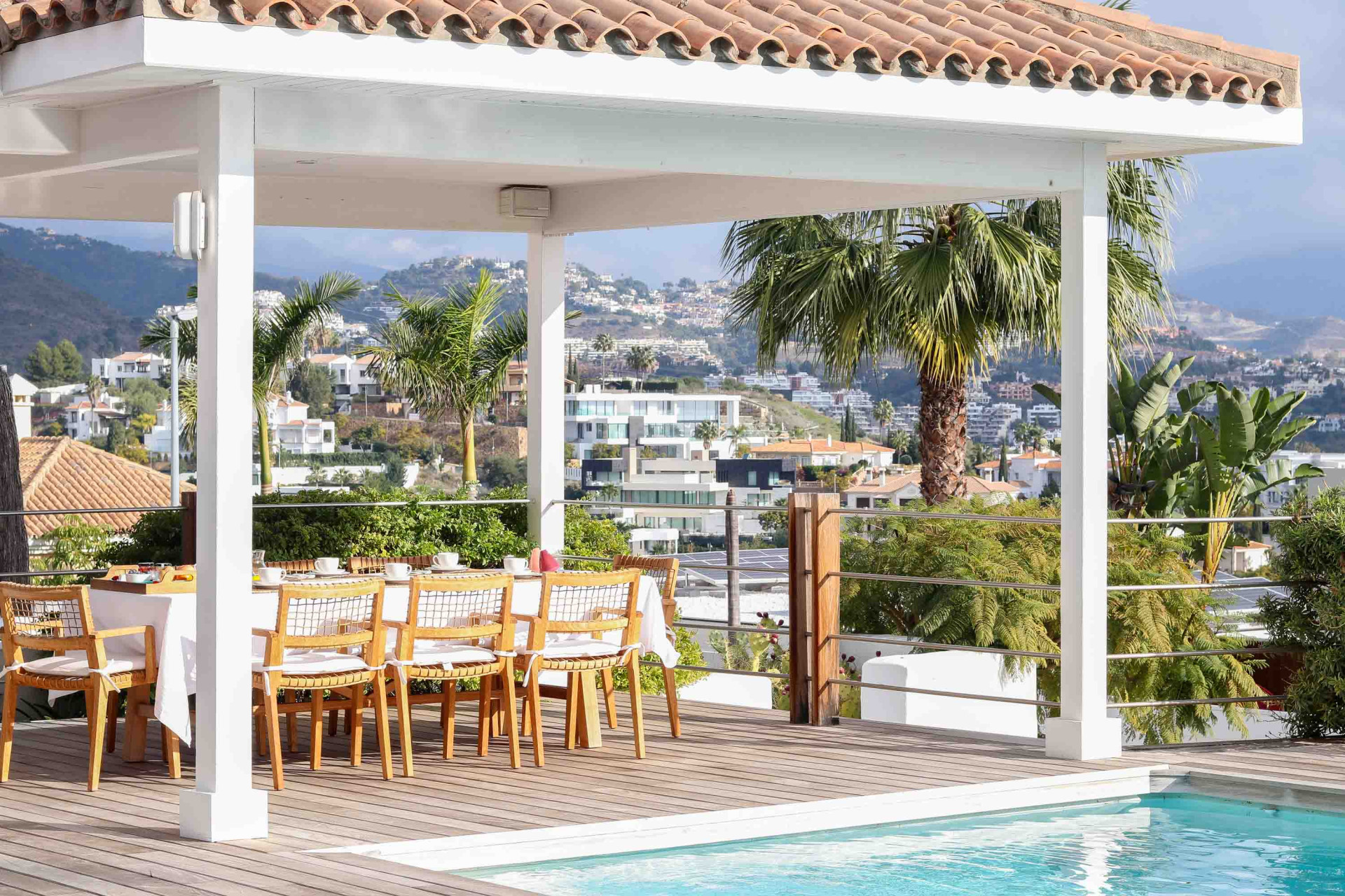 Outdoor covered dining area with a pool in the foreground and a panoramic view of distant hills and houses.