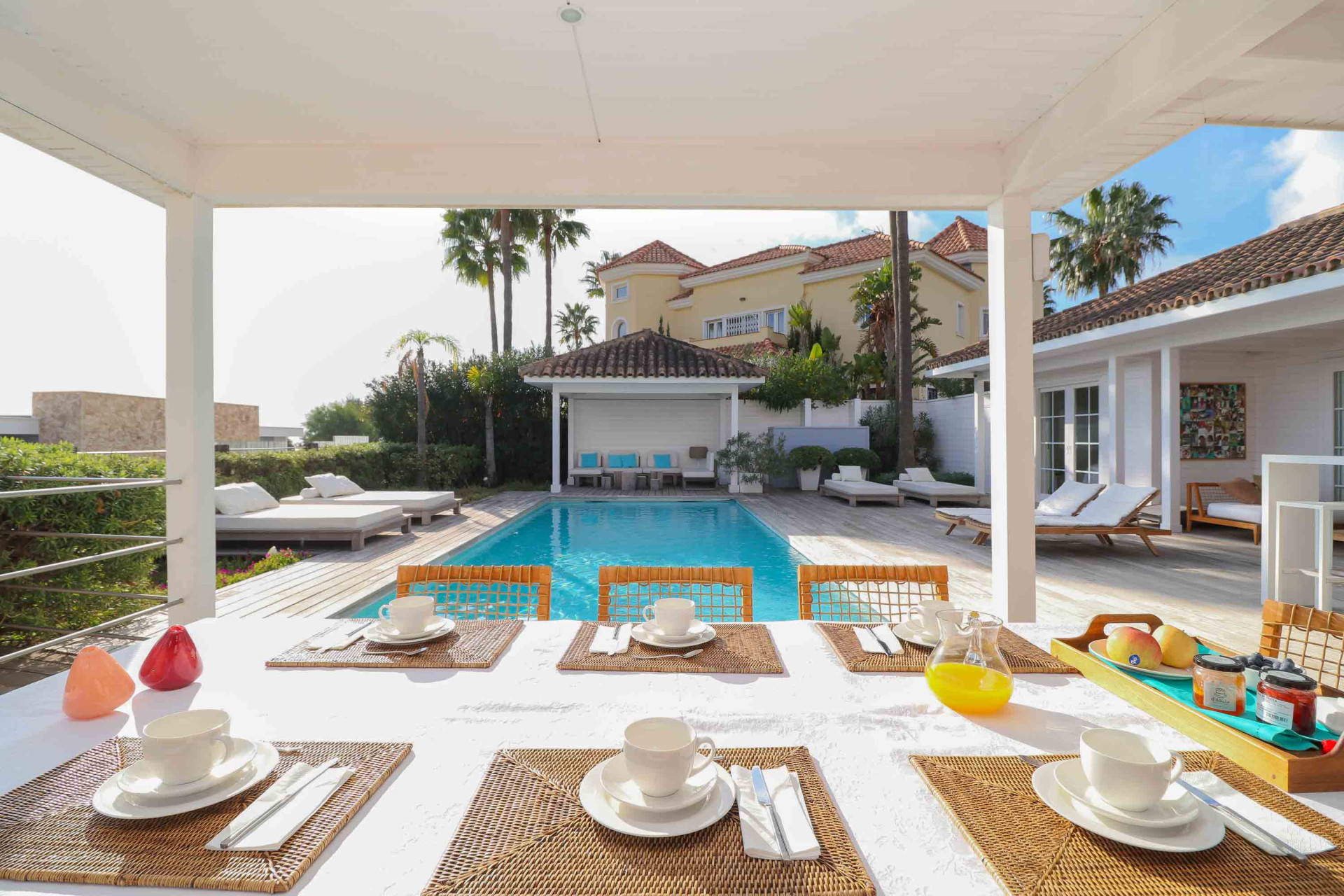 Outdoor dining table set under a covered patio, facing a swimming pool and a yellow villa.