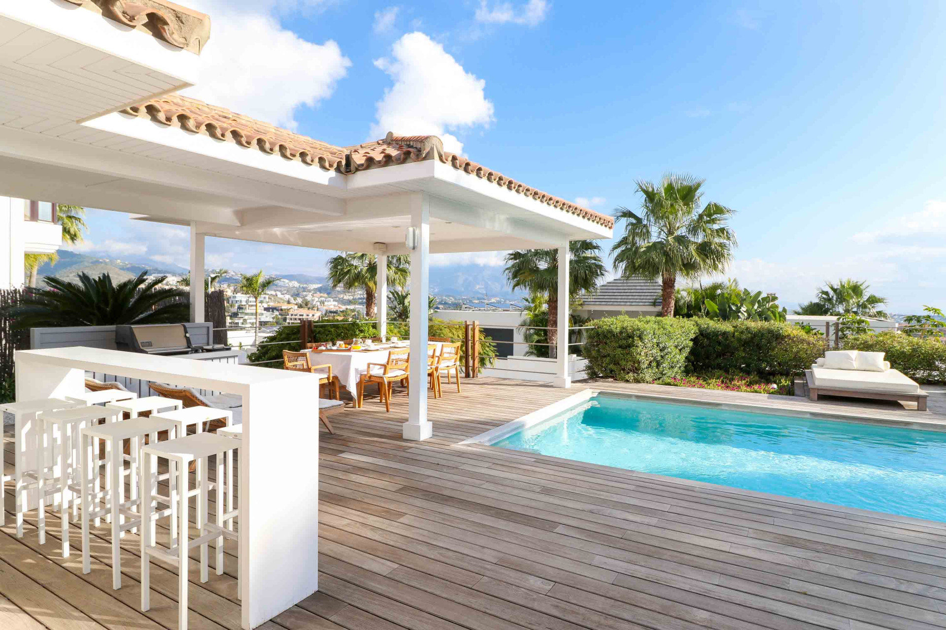 Outdoor patio with dining table, bar area, swimming pool, and mountain views under a sunny sky.