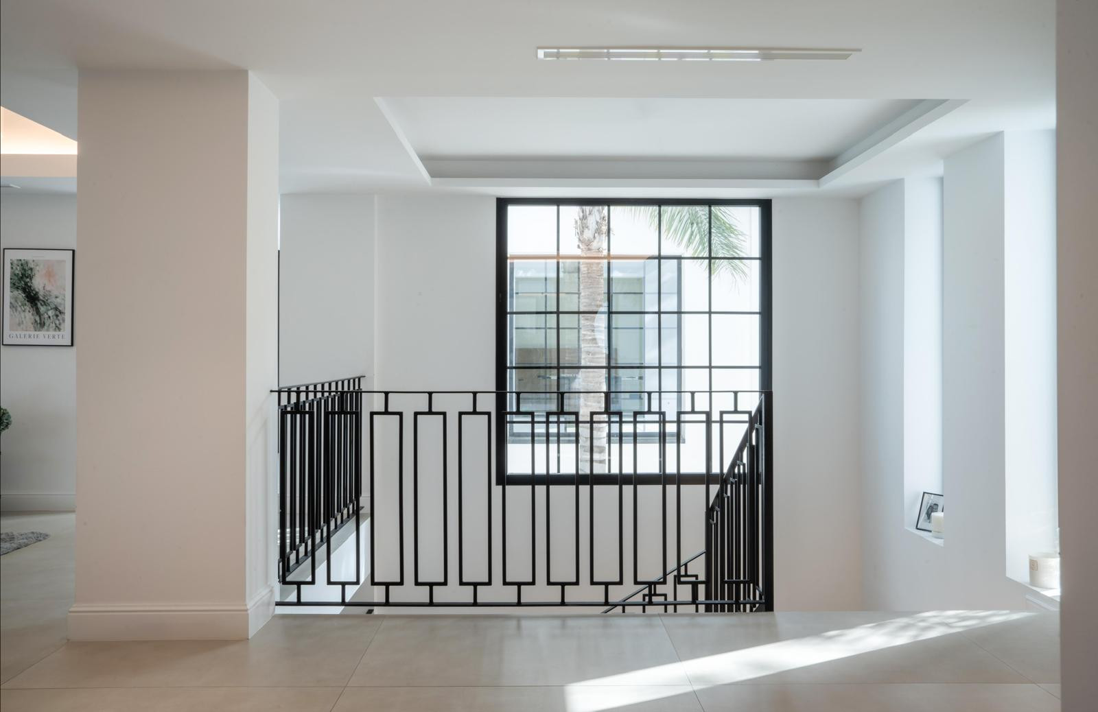 View of an open hallway with black railing and large window showing a palm tree outside.