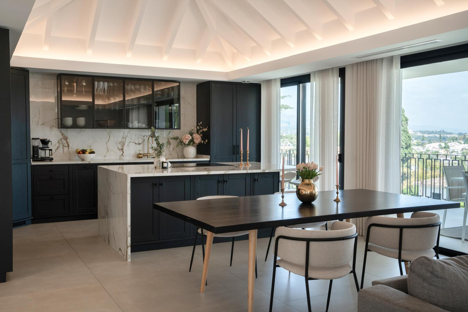 Modern kitchen with dark cabinets, marble countertops and large windows letting in natural light.