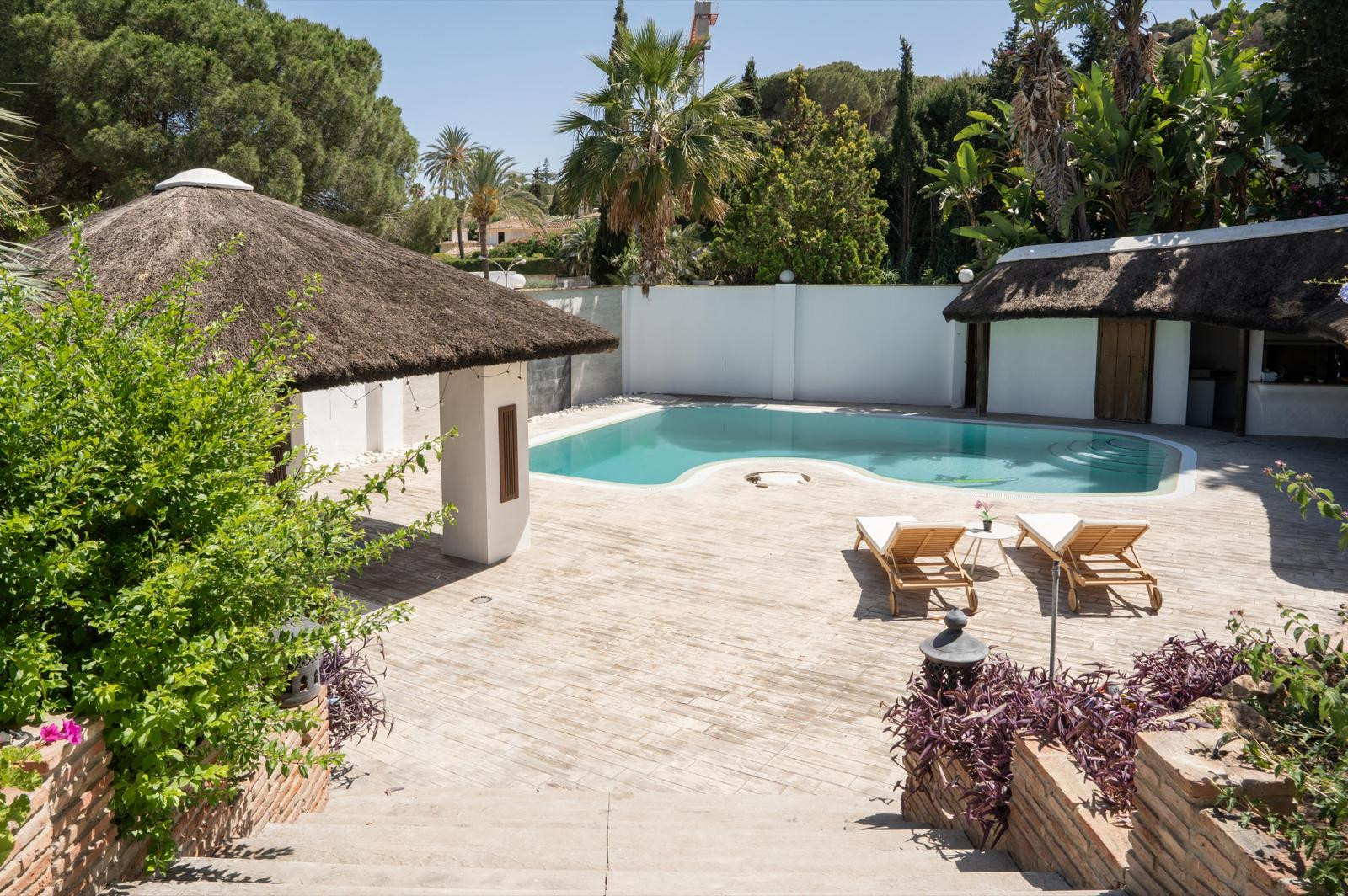 View of the pool area with a thatched roof gazebo and sun loungers surrounded by greenery.