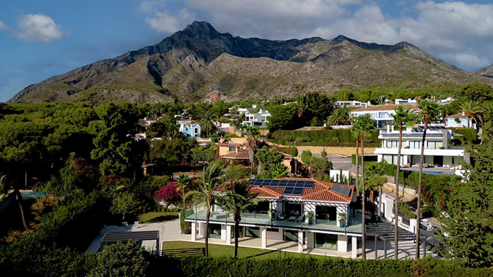 Aerial view of a white villa with a terracotta roof, solar panels, and glass railings, surrounded by lush trees and moun