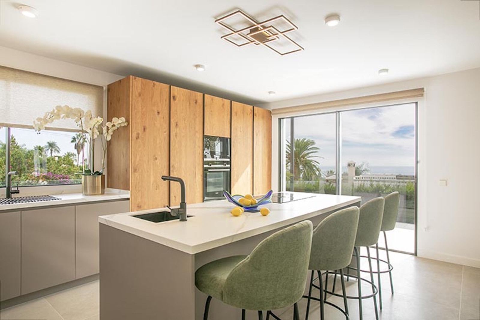 Modern kitchen with wood cabinets, white island with bar stools, and large window overlooking palm trees.