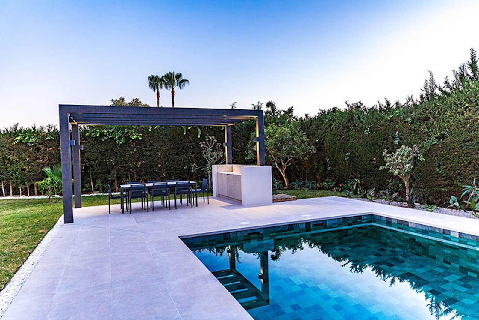 Outdoor pool and dining area with pergola and built-in barbecue station in a landscaped garden at dusk.