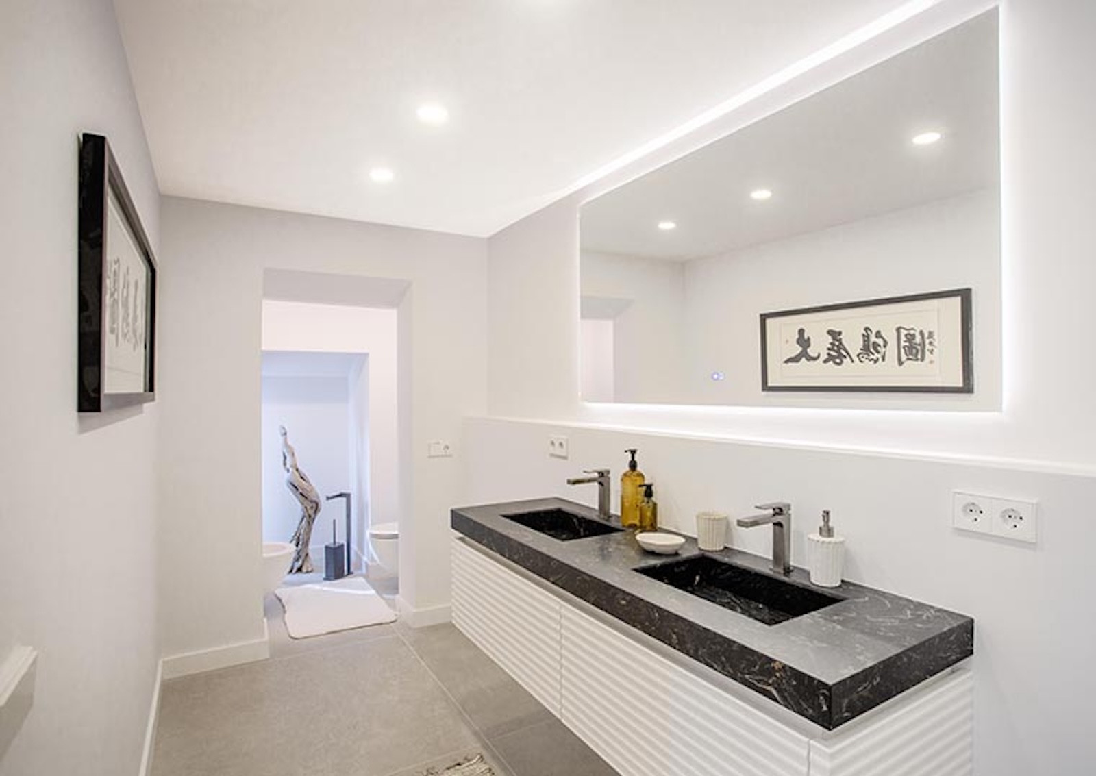 Bathroom with dual sinks, backlit mirror, and view into another area with a toilet and artwork.