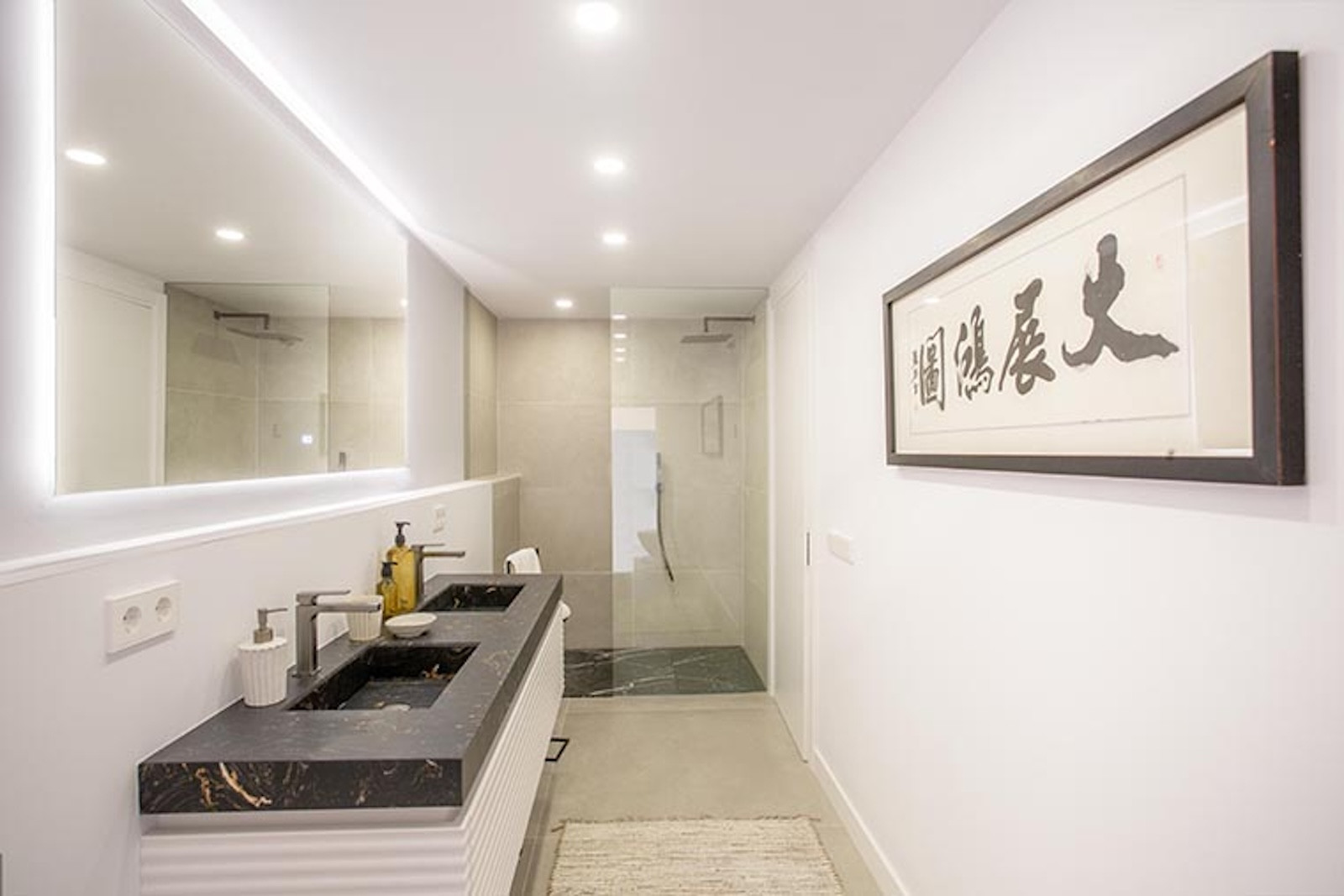 Bathroom with double black sinks, illuminated mirror, and a walk-in shower with dark floor tiles.