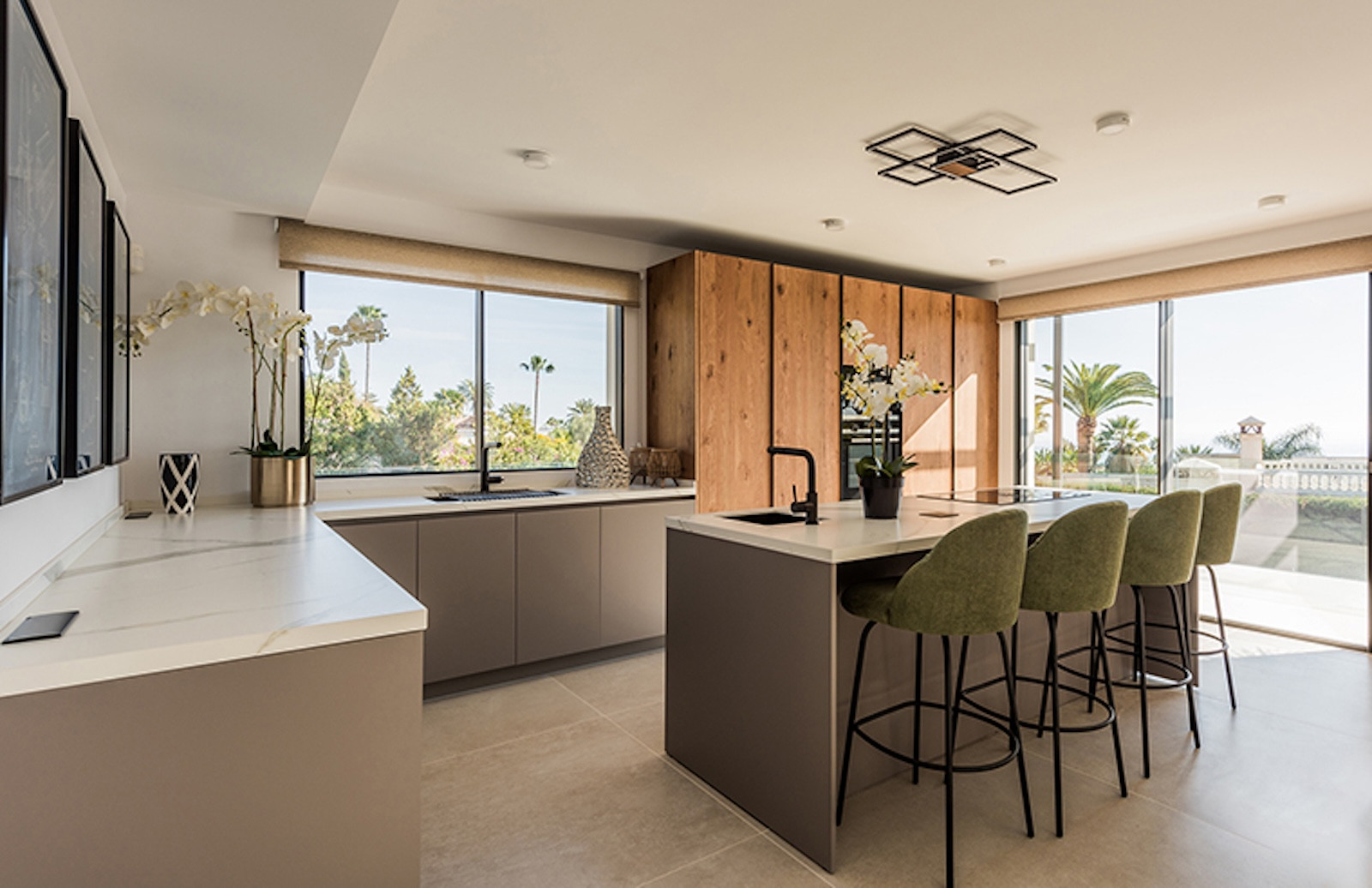 Modern kitchen with a white island, four olive green bar stools, and large windows revealing an outdoor area.