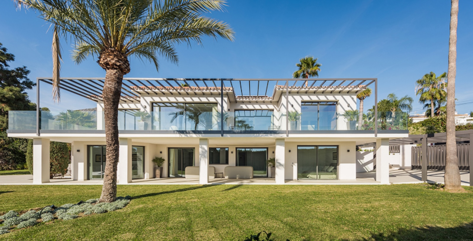 Front exterior of a white modern two-story villa with large windows and a manicured lawn under sunlight.