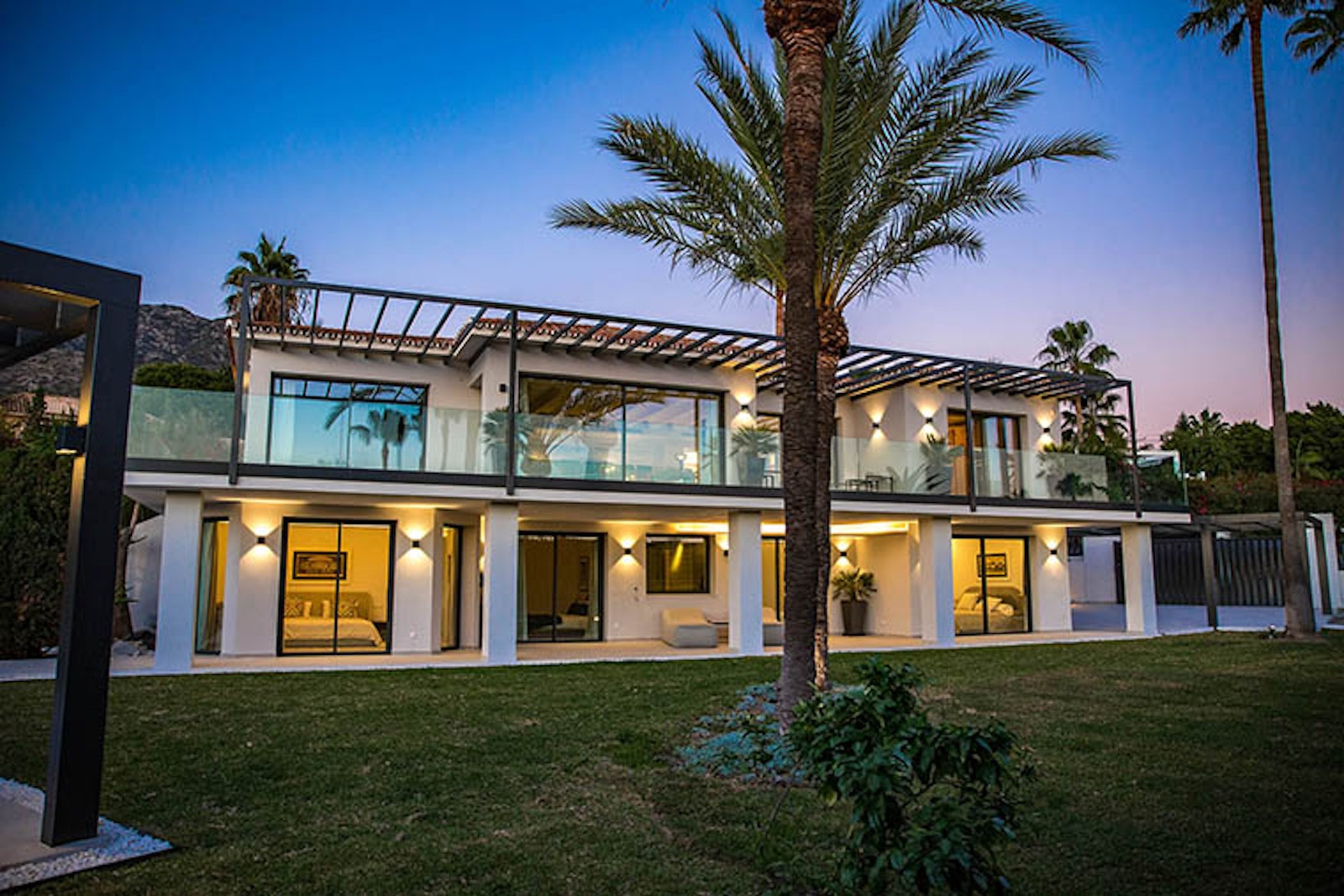 View of the brightly lit modern two-story villa from the lawn, showing balconies and large windows.