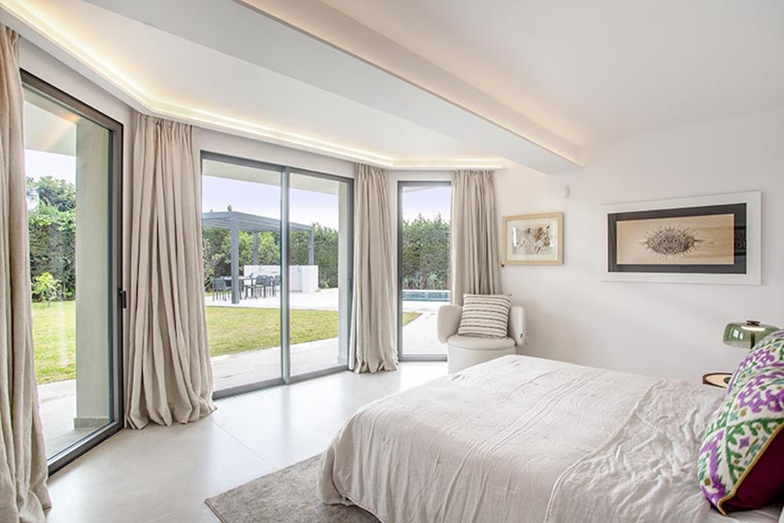 Bedroom with a large bed, armchair, and curved floor-to-ceiling glass doors opening to a lawn.