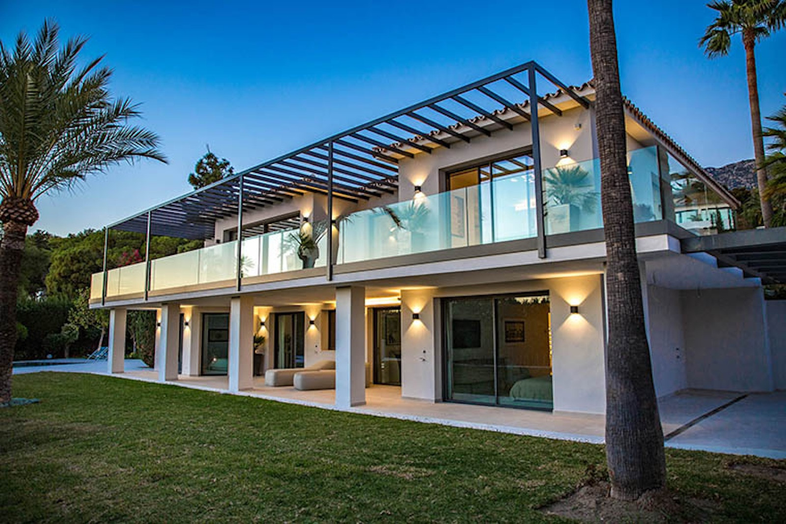 Exterior of a modern two-story villa at dusk, with glass railings, outdoor lighting, and palm trees.