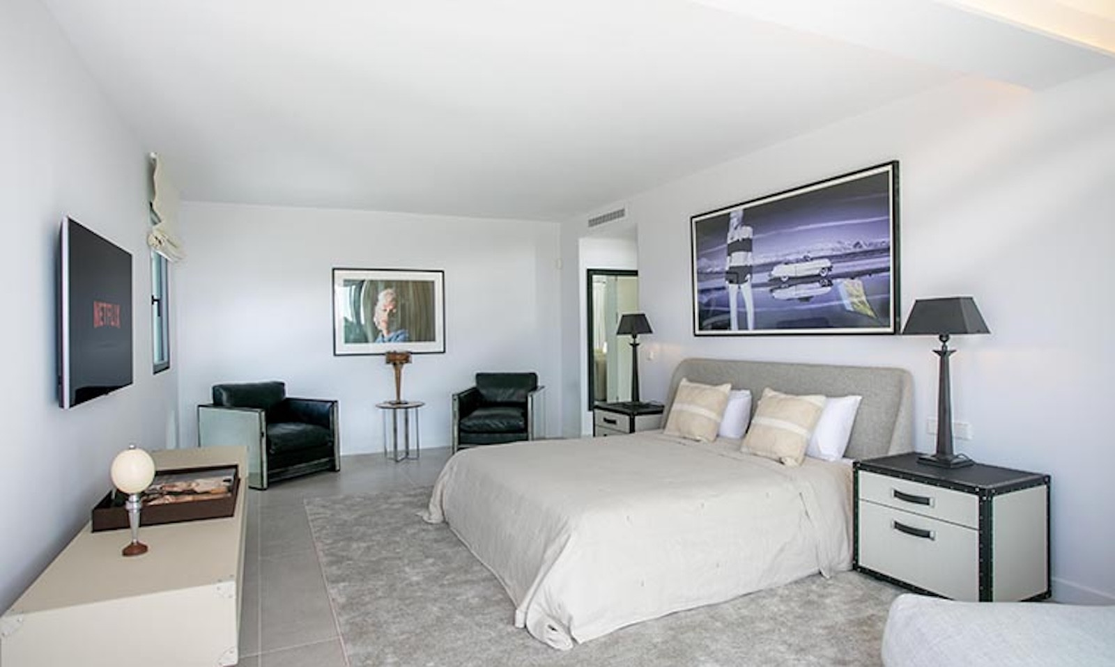 Bedroom with a bed, two dark side tables, two black armchairs, a wall-mounted TV, and artwork.