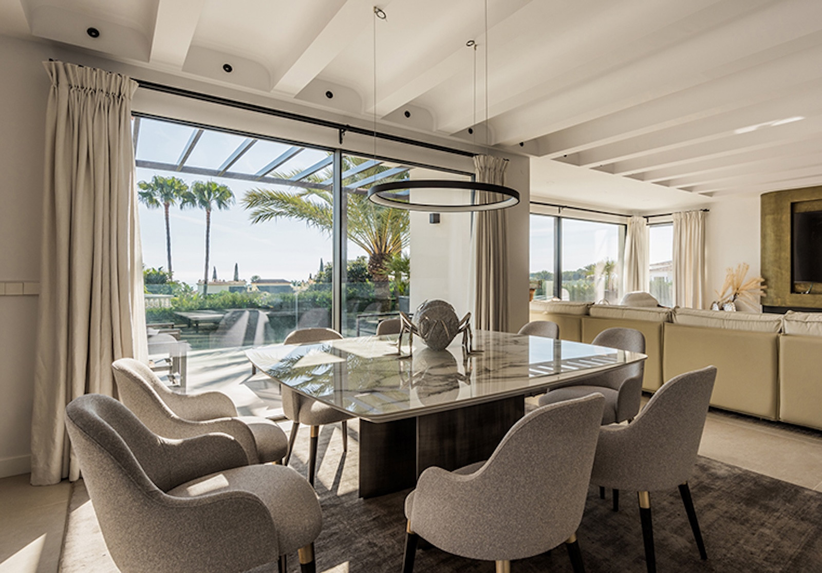 Dining room with six grey chairs around a marble table, palm trees visible through large windows.