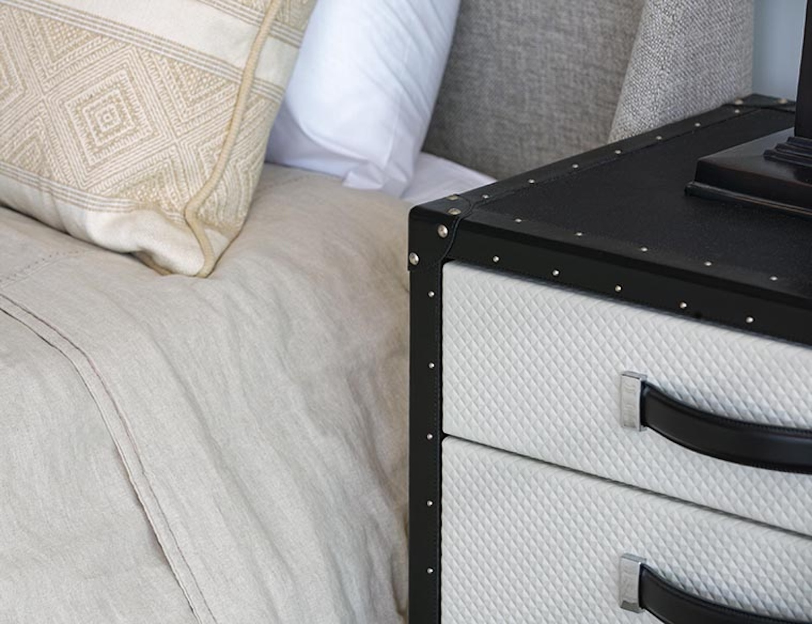 Close up of a bed with light bedding next to a black and white trunk style nightstand.