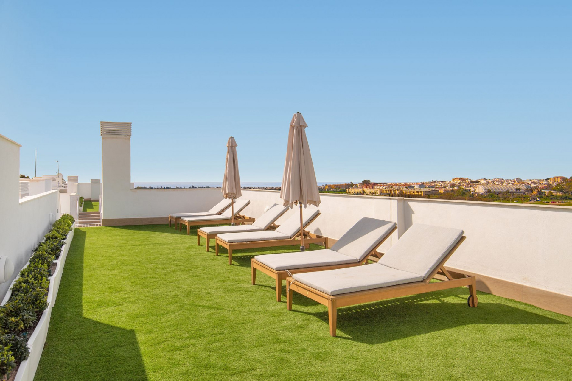 Rooftop lounge area with four wooden sun loungers, three open umbrellas, and artificial grass.