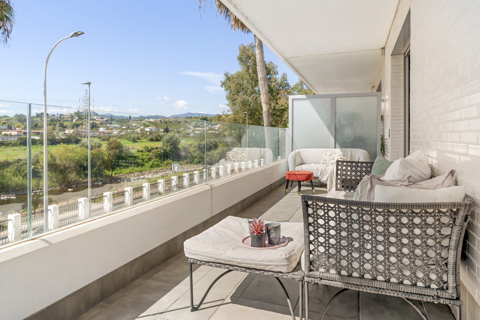 Balcony with a sofa and armchair, facing a glass railing with views of mountains and a river.