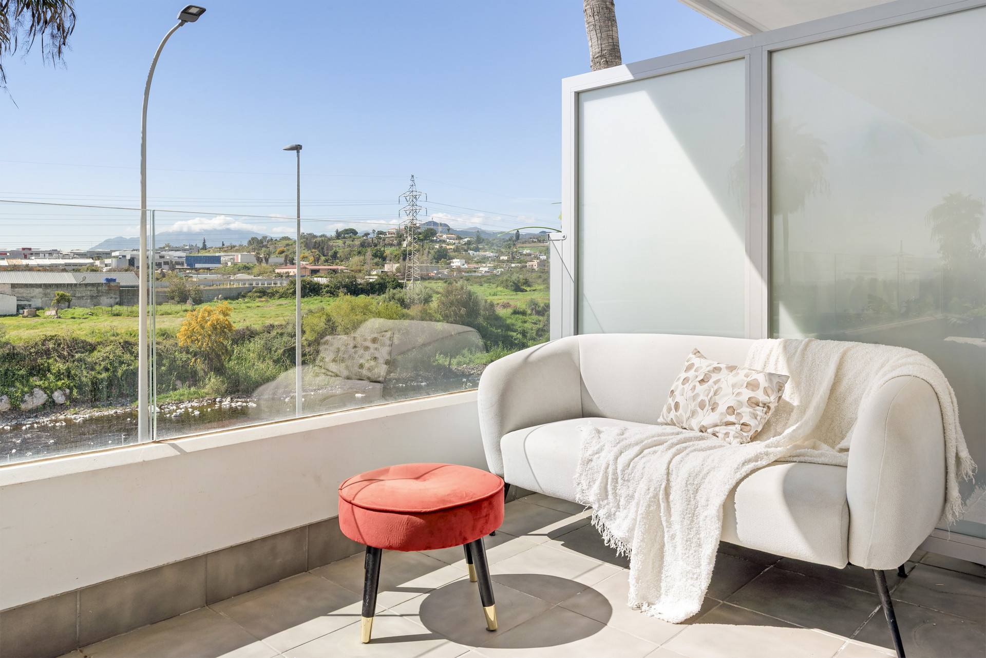 Balcony nook with a light gray sofa, red ottoman, and a panoramic view of the landscape.
