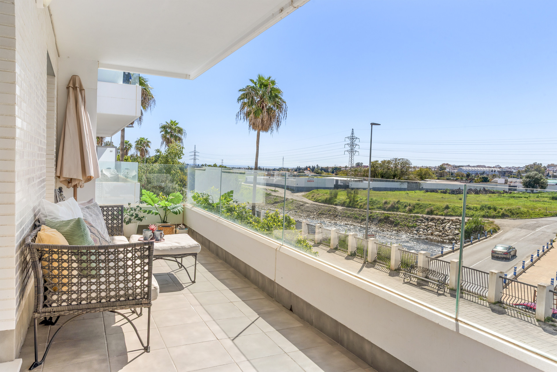 Balcony with outdoor seating, a glass railing, and a view of palm trees and a river.