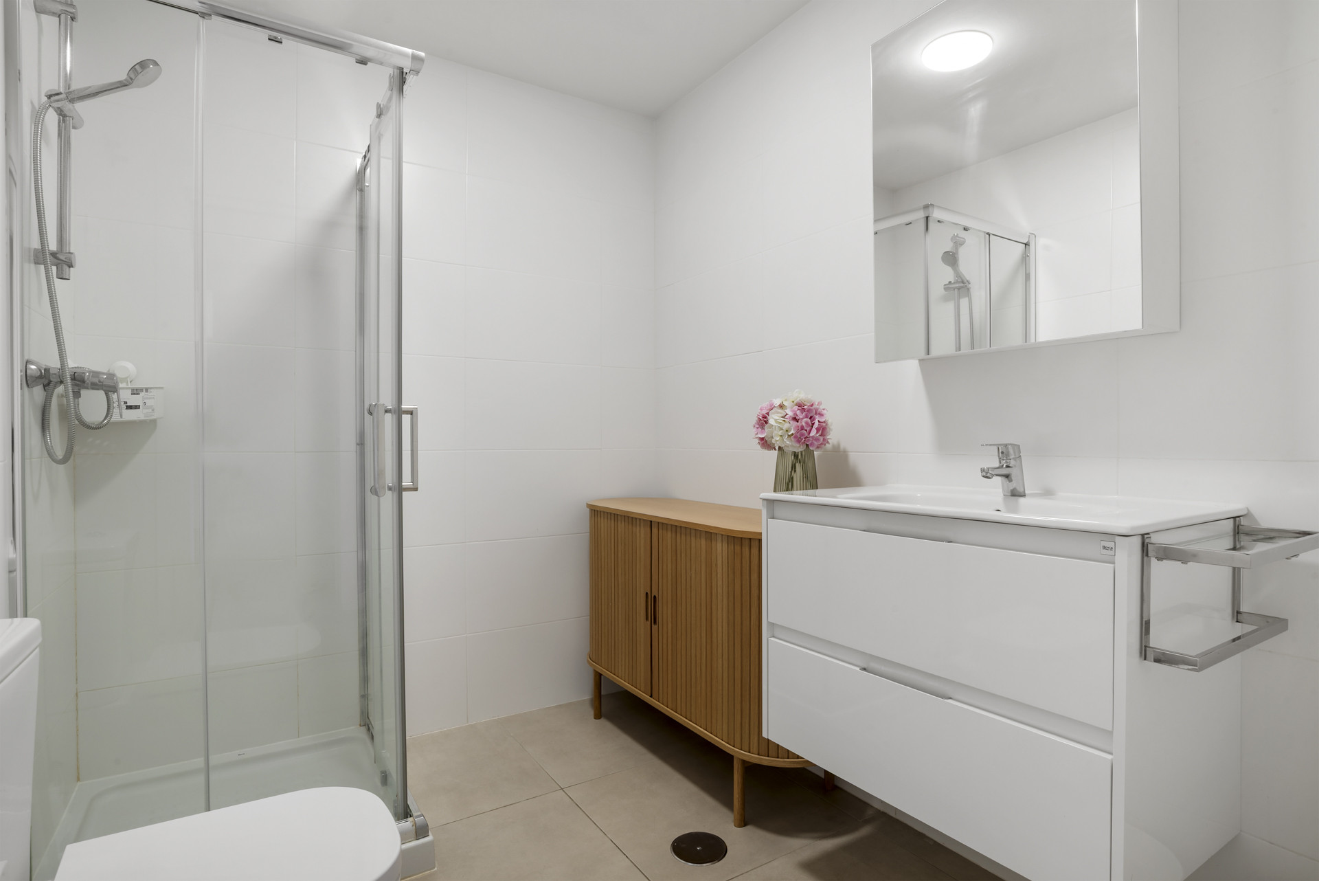 White tiled bathroom with a glass shower, toilet, modern white vanity, and small wooden cabinet.