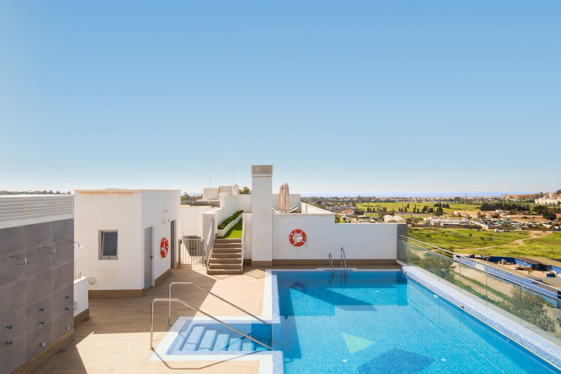 Rooftop swimming pool with blue tiles, surrounded by white walls and a view of the landscape.