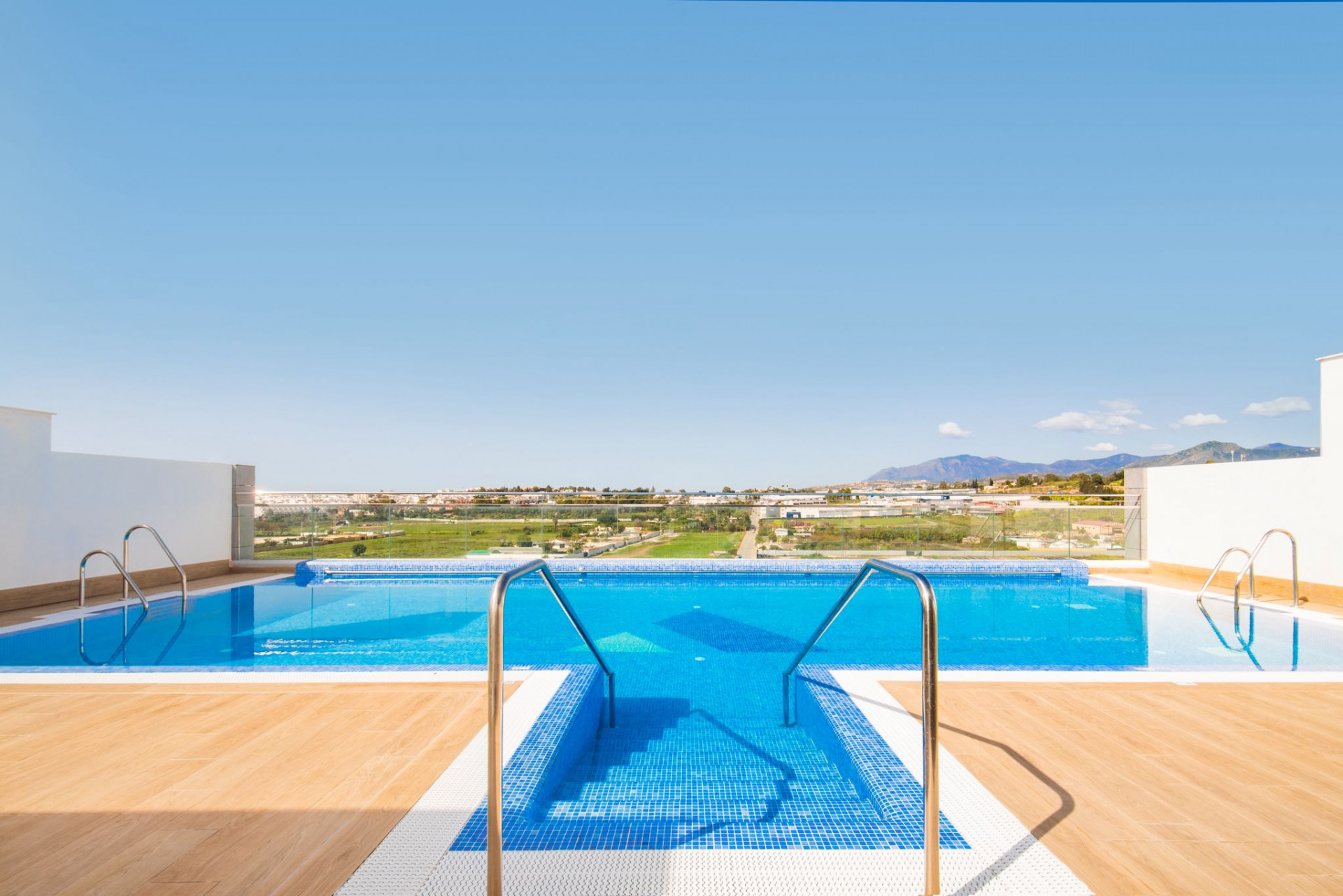 Outdoor rooftop pool area with blue tiled pools, wooden decking, and a distant city and mountain view.