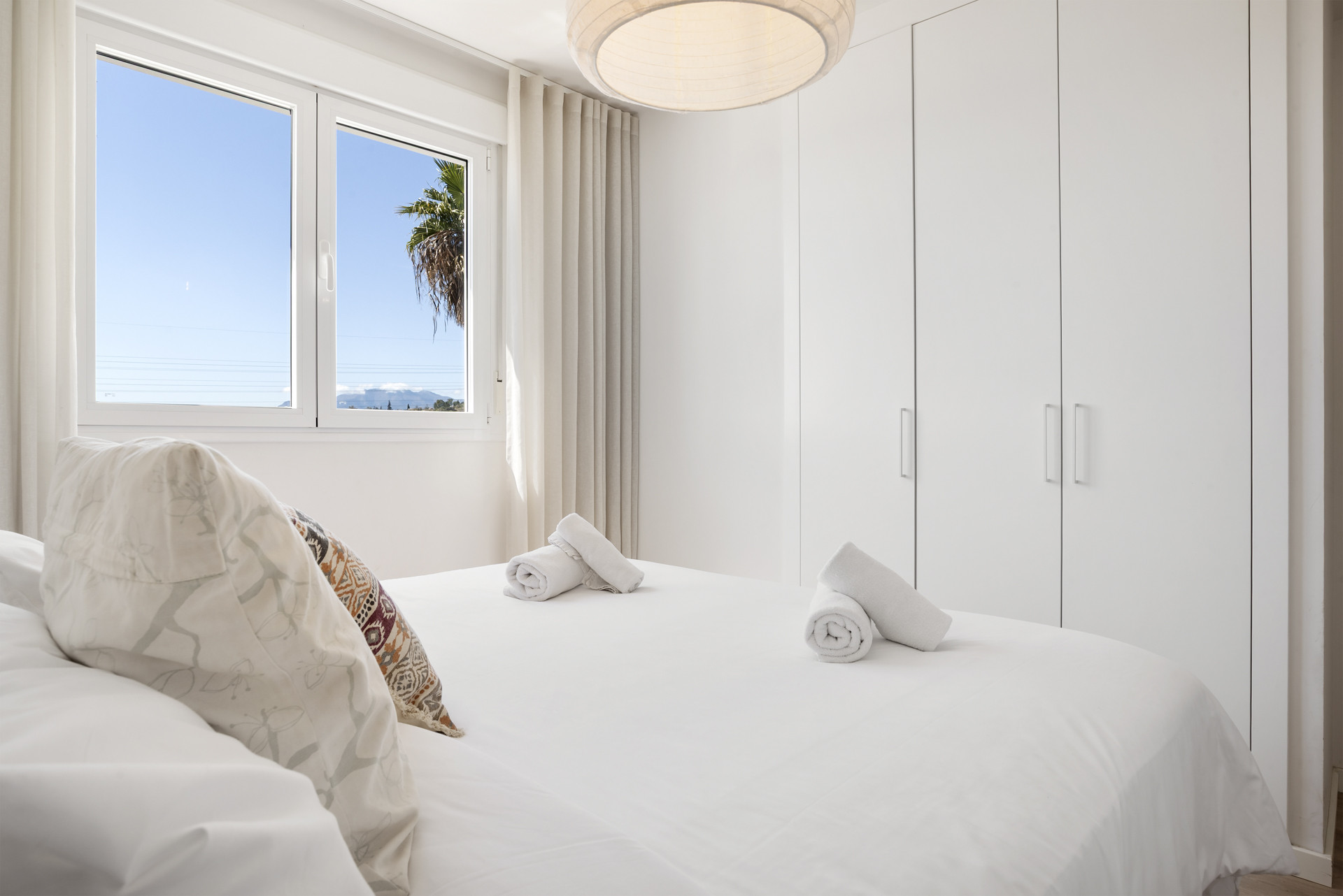 Bedroom with white bed linens, rolled towels, white built-in wardrobes, and a window showing foliage.