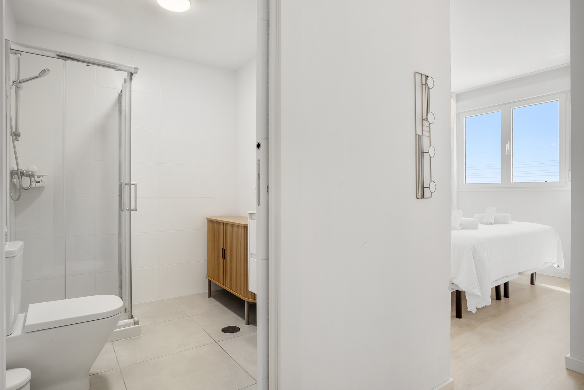 Bathroom with a shower, toilet, and wooden vanity, with a bedroom visible through the doorway.