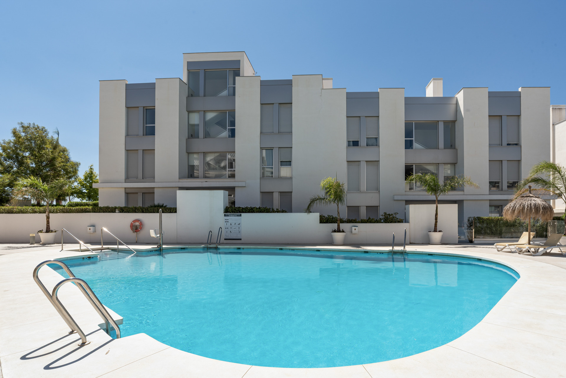 Swimming pool with metal handrails in front of a multi-story residential building under a clear sky.