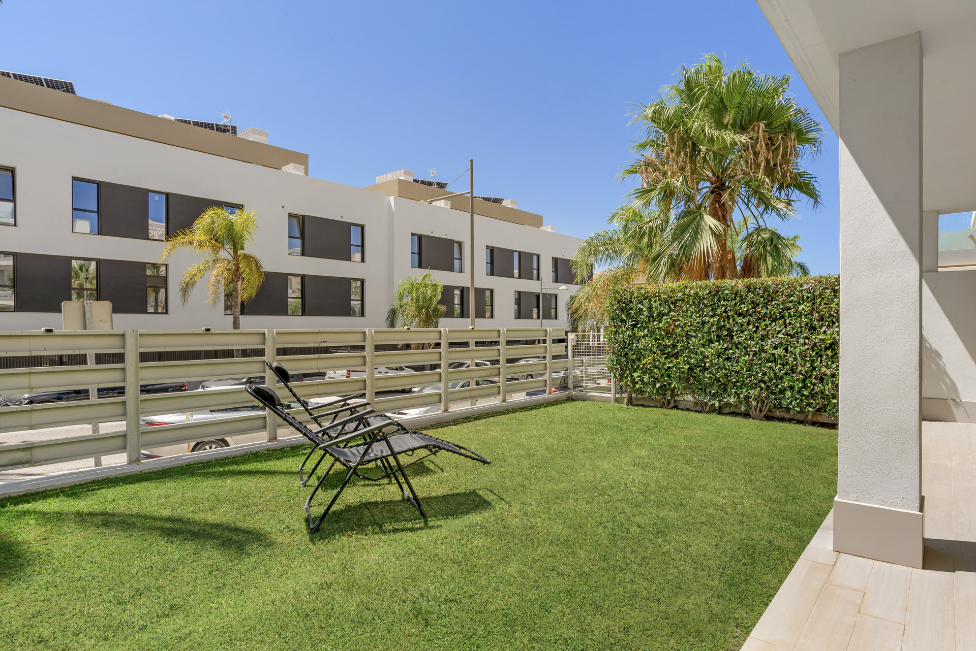 Lawn area with two black lounge chairs, a low fence, and a row of buildings in the background.