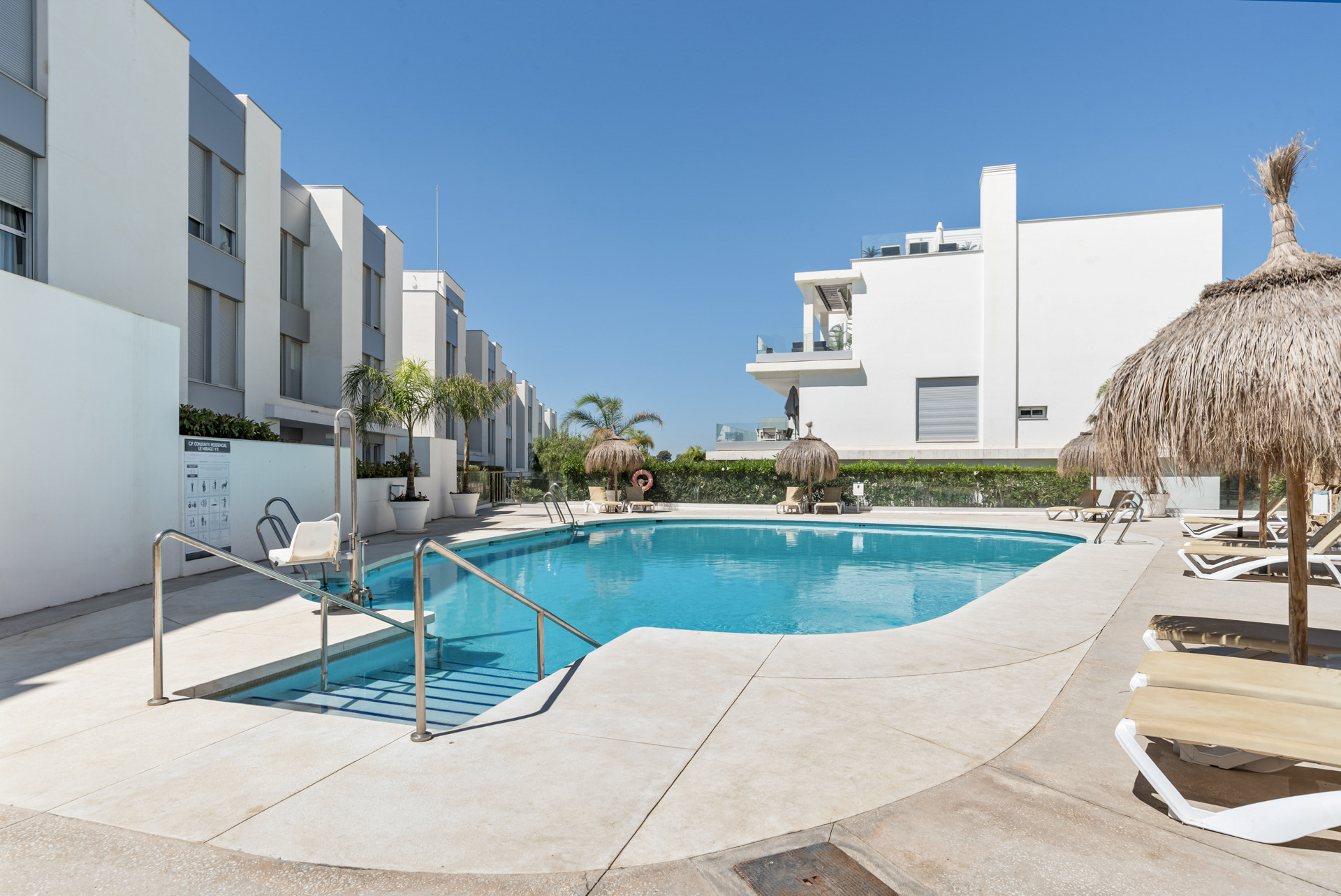Outdoor swimming pool surrounded by modern white buildings, straw umbrellas, and lounge chairs.