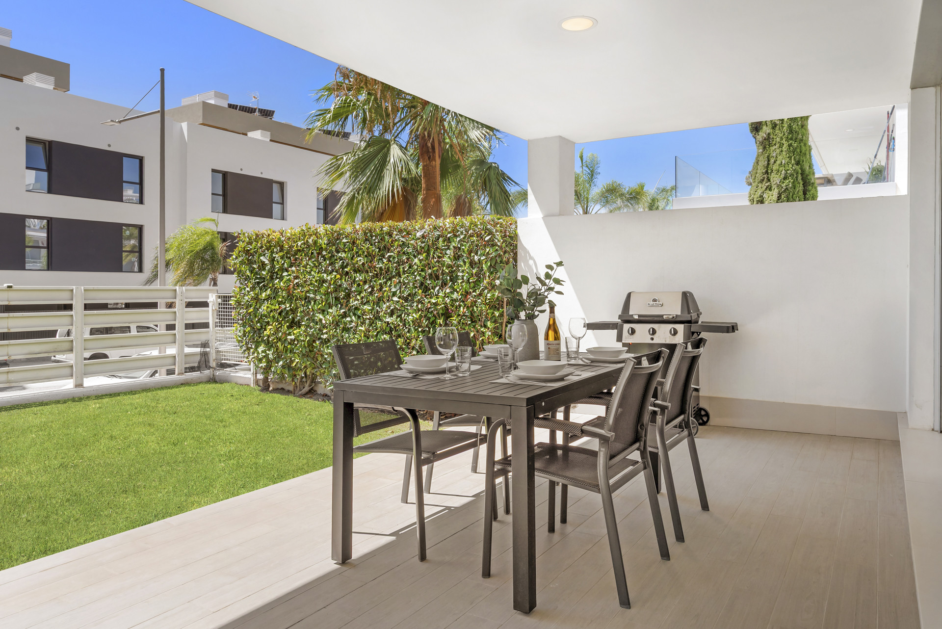 Covered outdoor patio with a dining table set for six, a barbecue grill, and a green lawn bordered by a hedge.