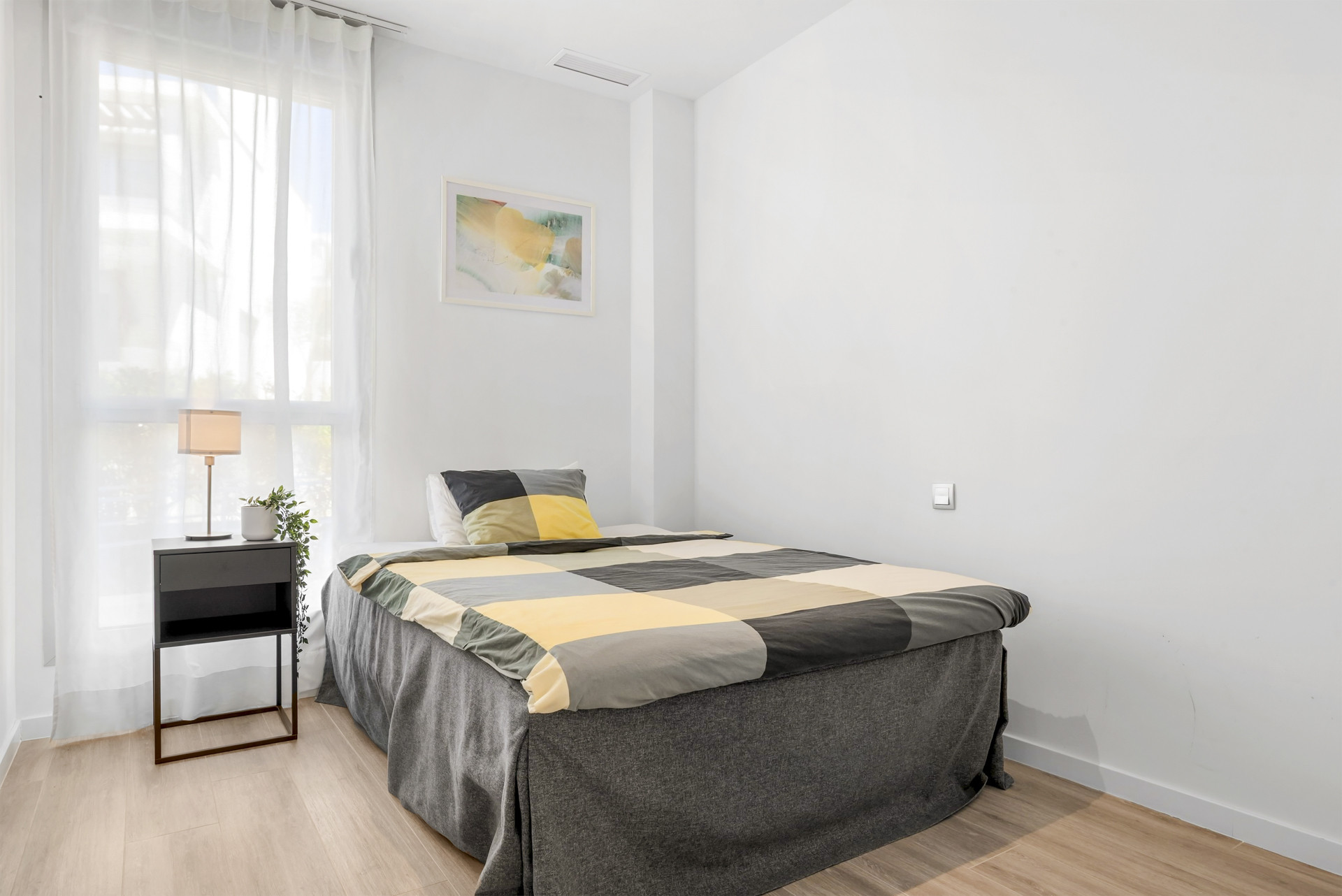 Bedroom with a large window, light wood floor, and a bed with a gray and yellow checkered duvet.