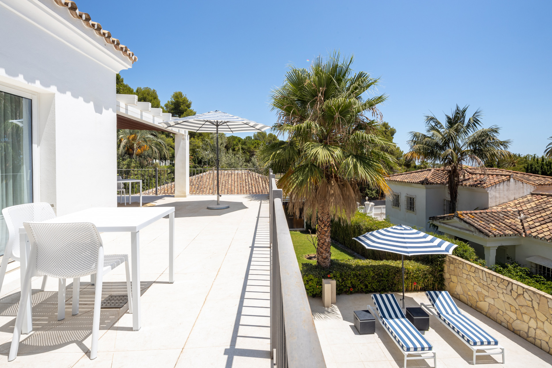 Large white rooftop terrace with striped umbrella, lounge chairs, a table, and palm trees.