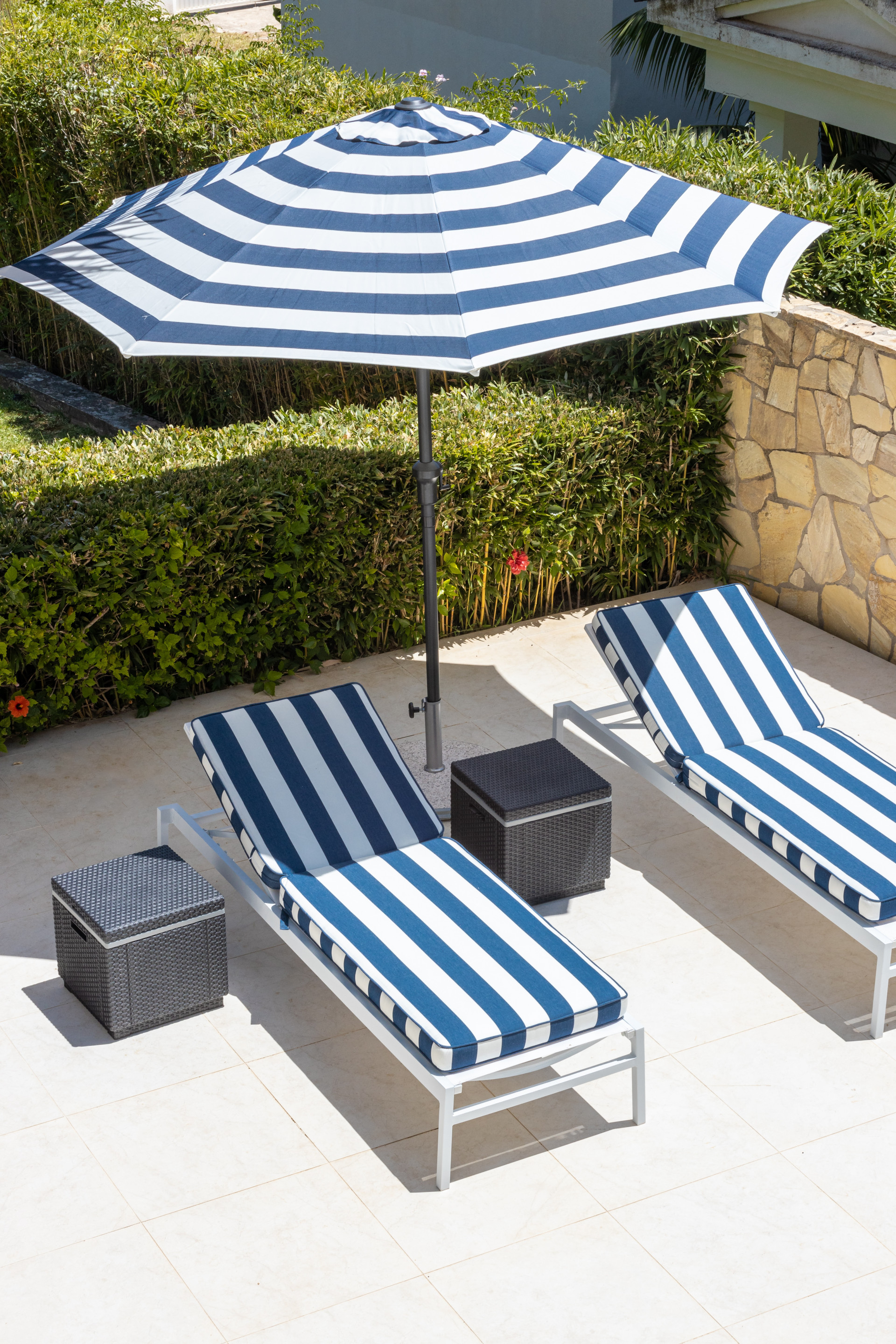 Two striped blue and white lounge chairs and a matching umbrella on a patio near a hedge.