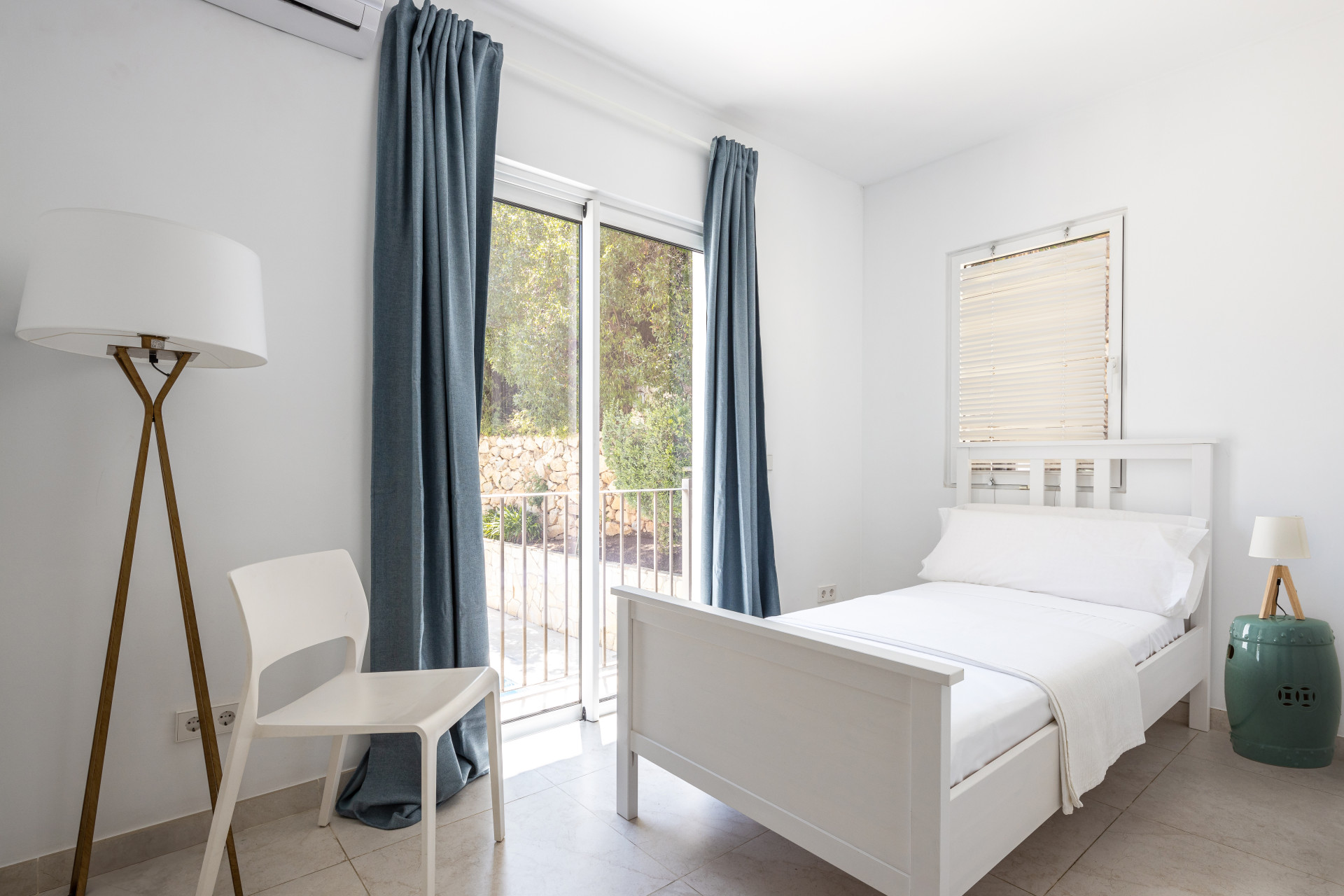 Bedroom with a single white bed, floor lamp, white chair, and glass door overlooking a patio.