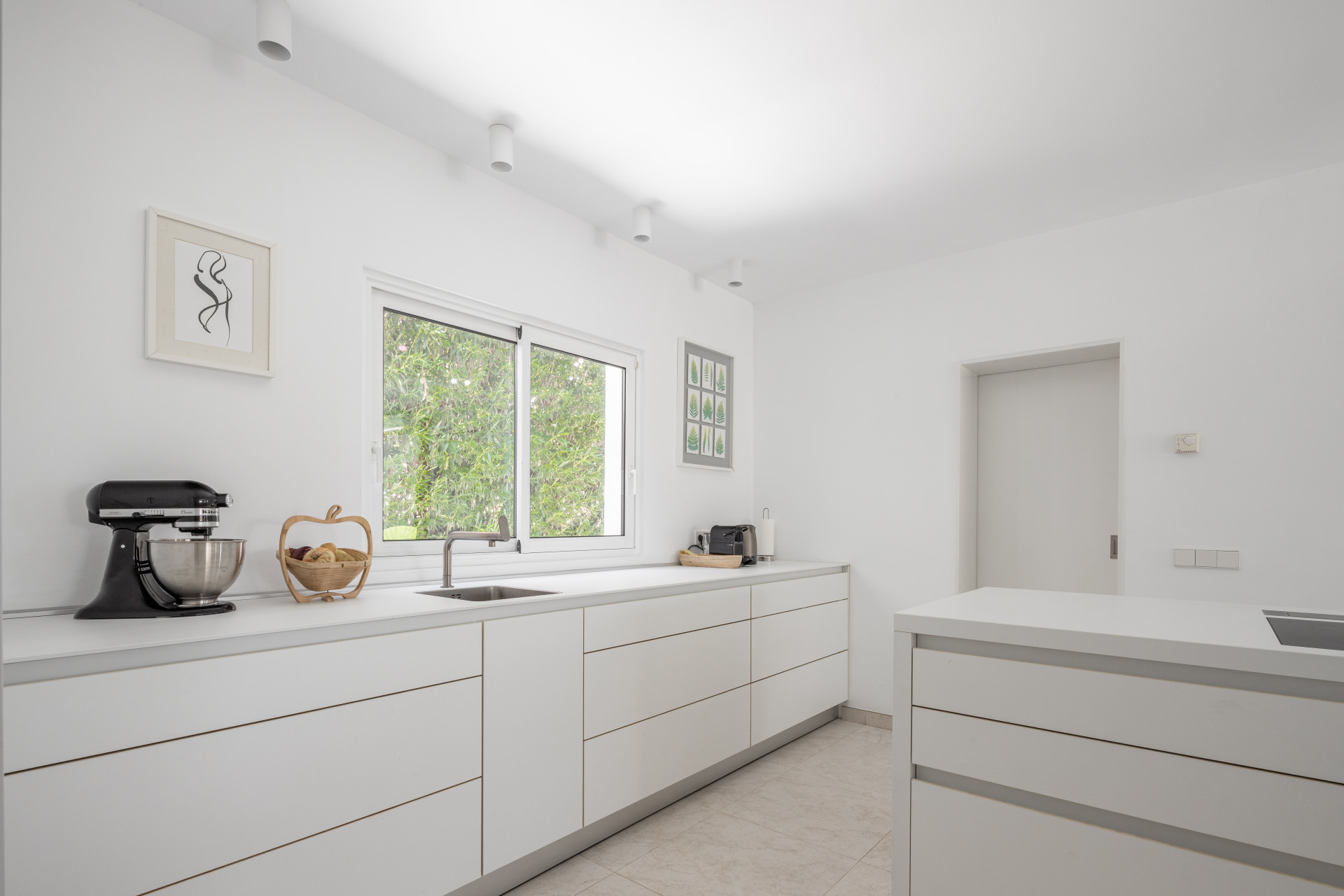 Modern white kitchen with stainless steel sink, large window, and light tile flooring.