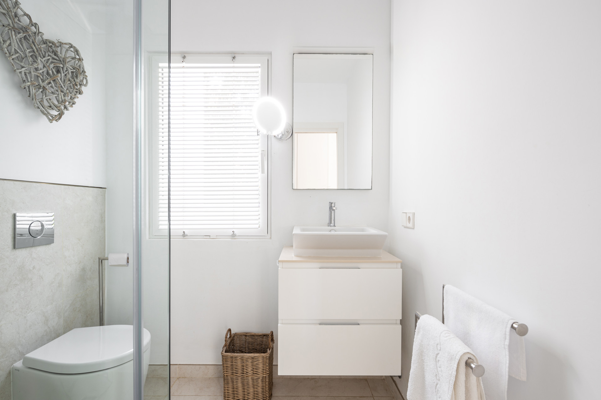 White bathroom with toilet, glass shower, and vanity with rectangular sink and round lighted mirror.