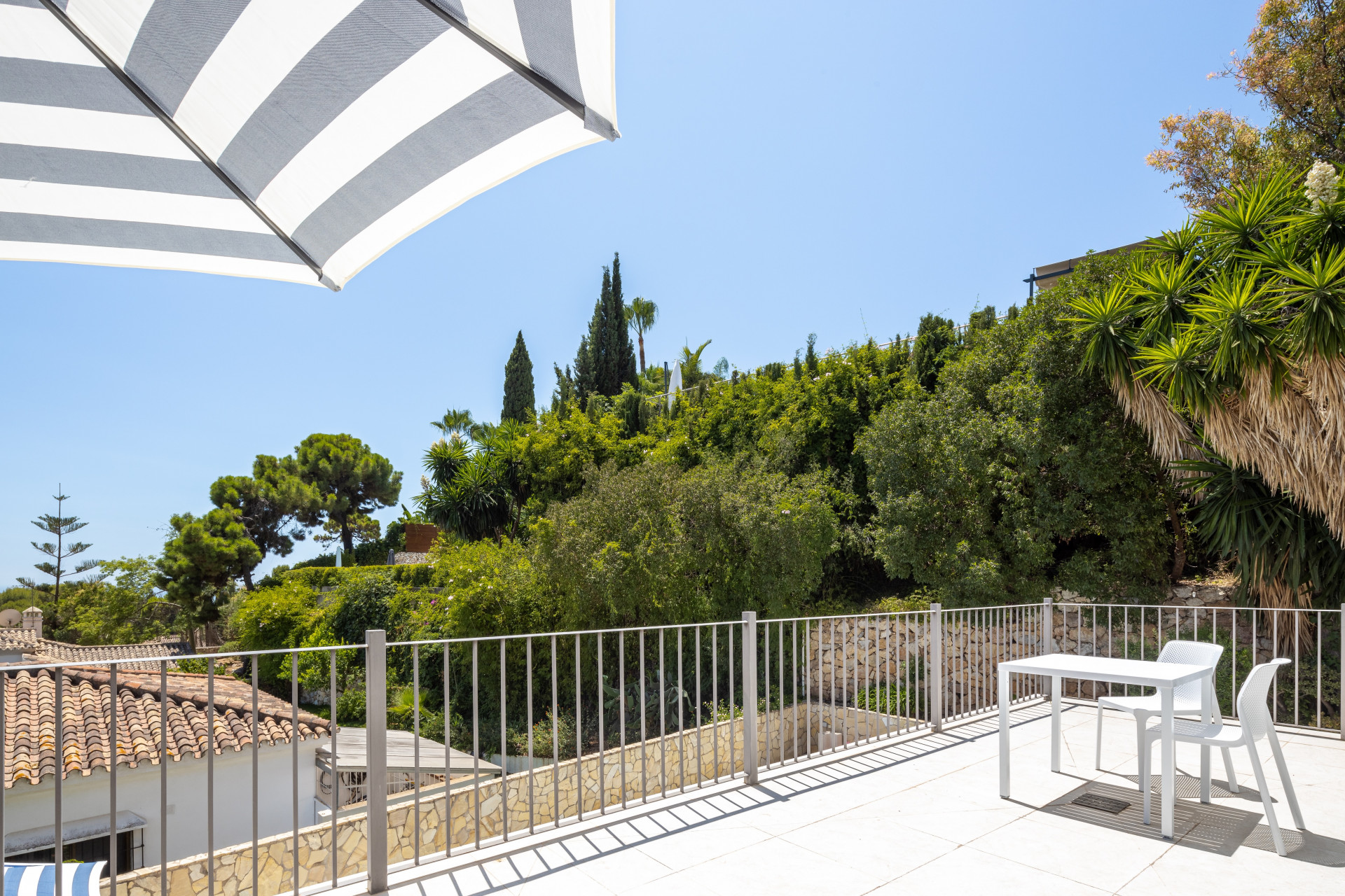Rooftop terrace with white table and chairs, a striped umbrella, overlooking green hills and blue sky.