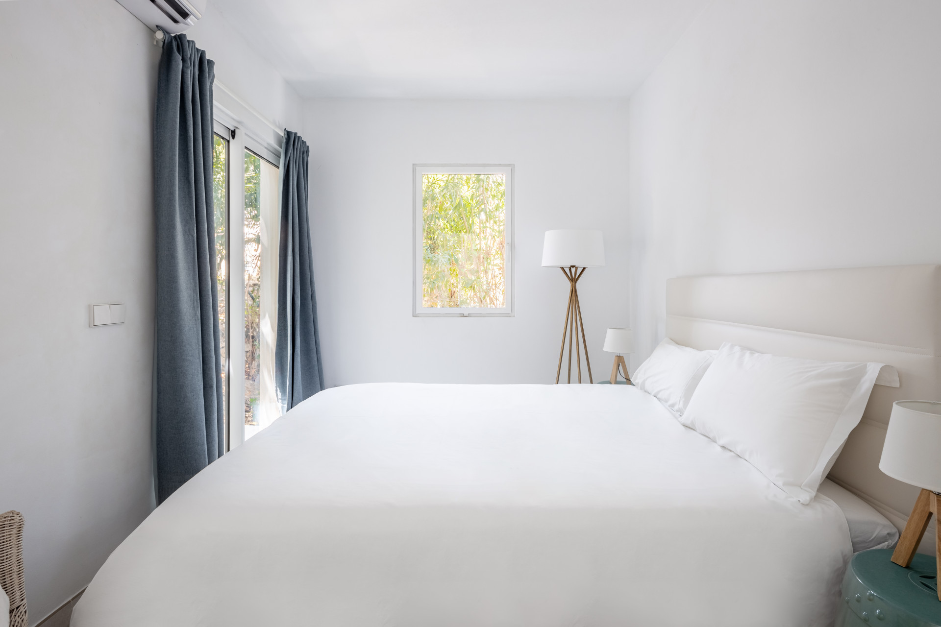 Bedroom with a large white bed, two lamps, a small window, and a glass door with blue curtains.