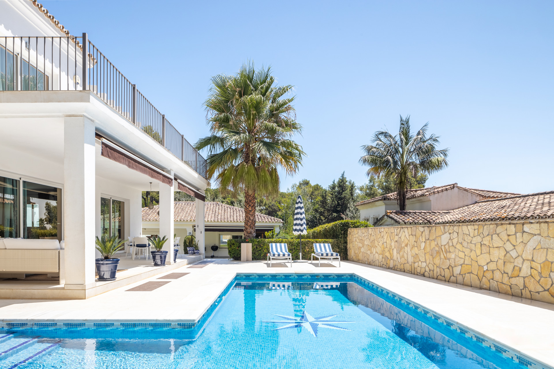 Exterior view of villa with rectangular pool, patio, palm tree, and clear blue sky.
