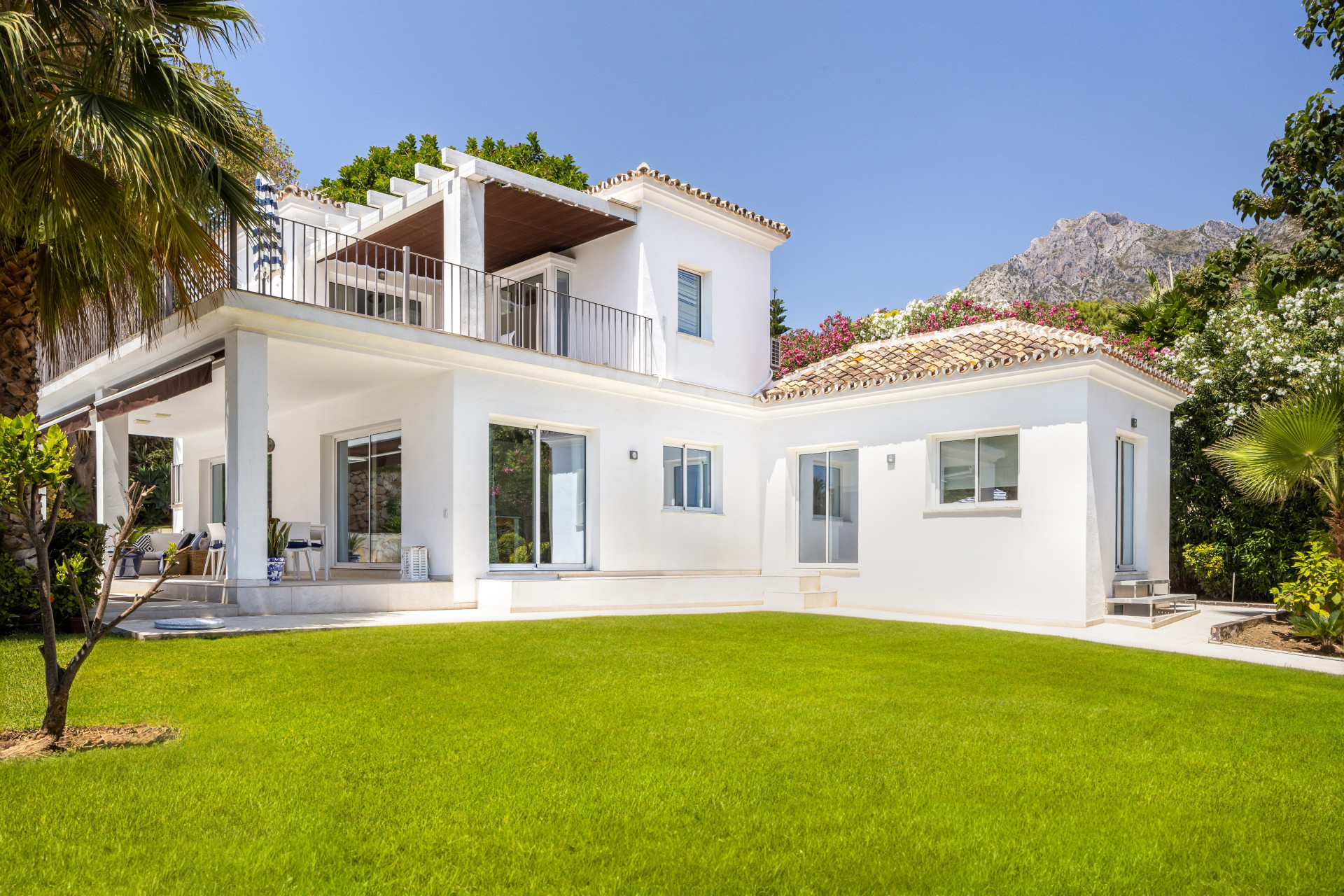 White two-story villa with tiled roof, large green lawn, and mountains in the background.