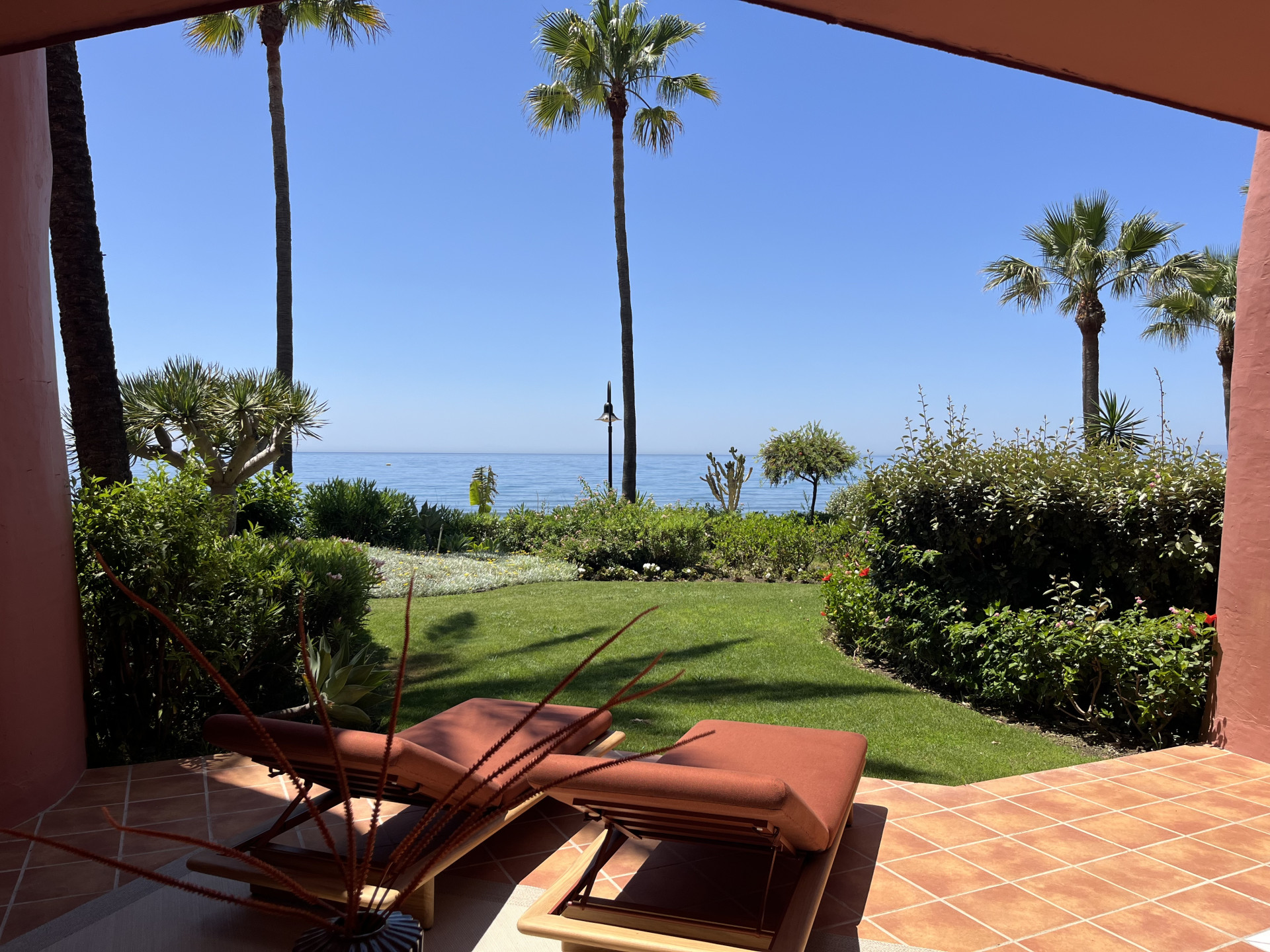 View from a patio with two lounge chairs, green lawn, palm trees, and the blue ocean in the distance.