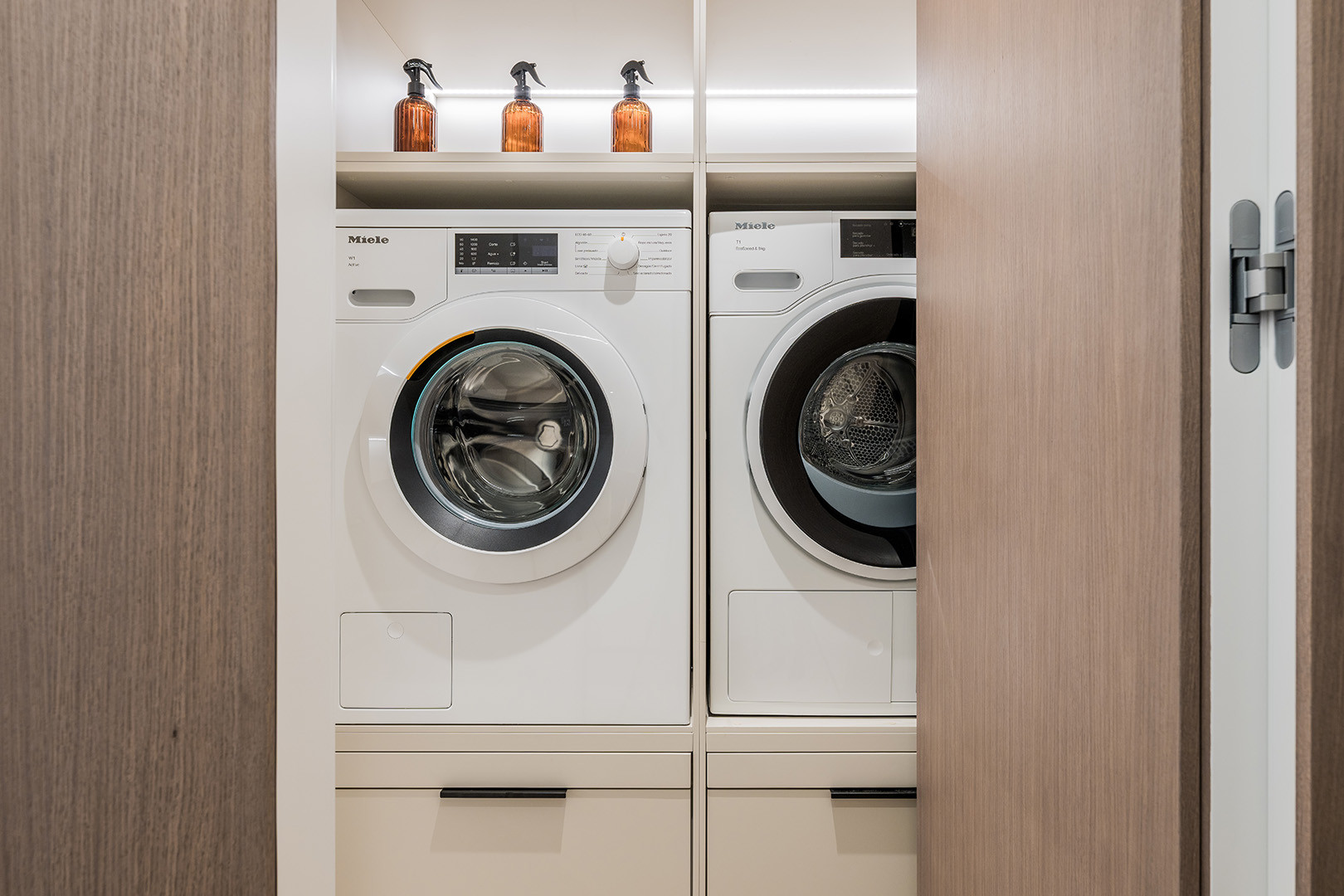 Laundry closet with stacked white Miele washer and dryer units, and amber spray bottles on a shelf.