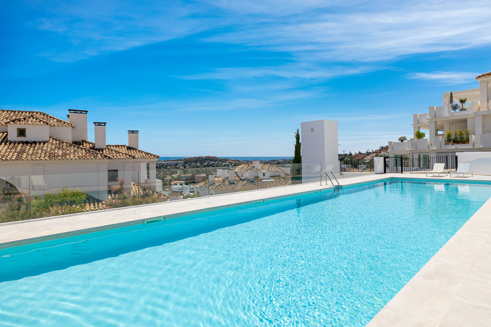 Outdoor infinity pool overlooking white residential buildings and distant hills under a blue sky.