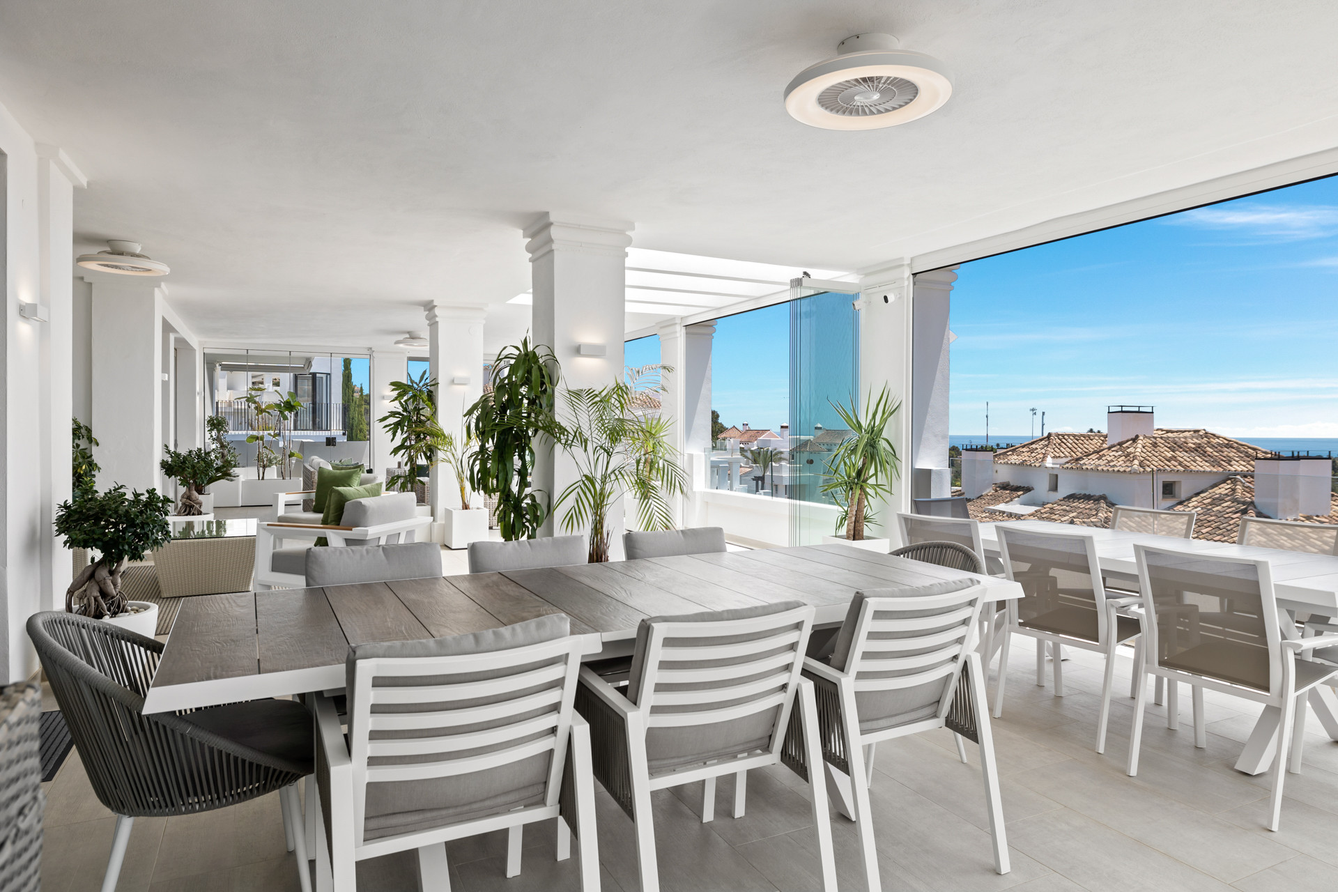 Covered outdoor dining area with two long tables, chairs, and large glass panels overlooking the sea.