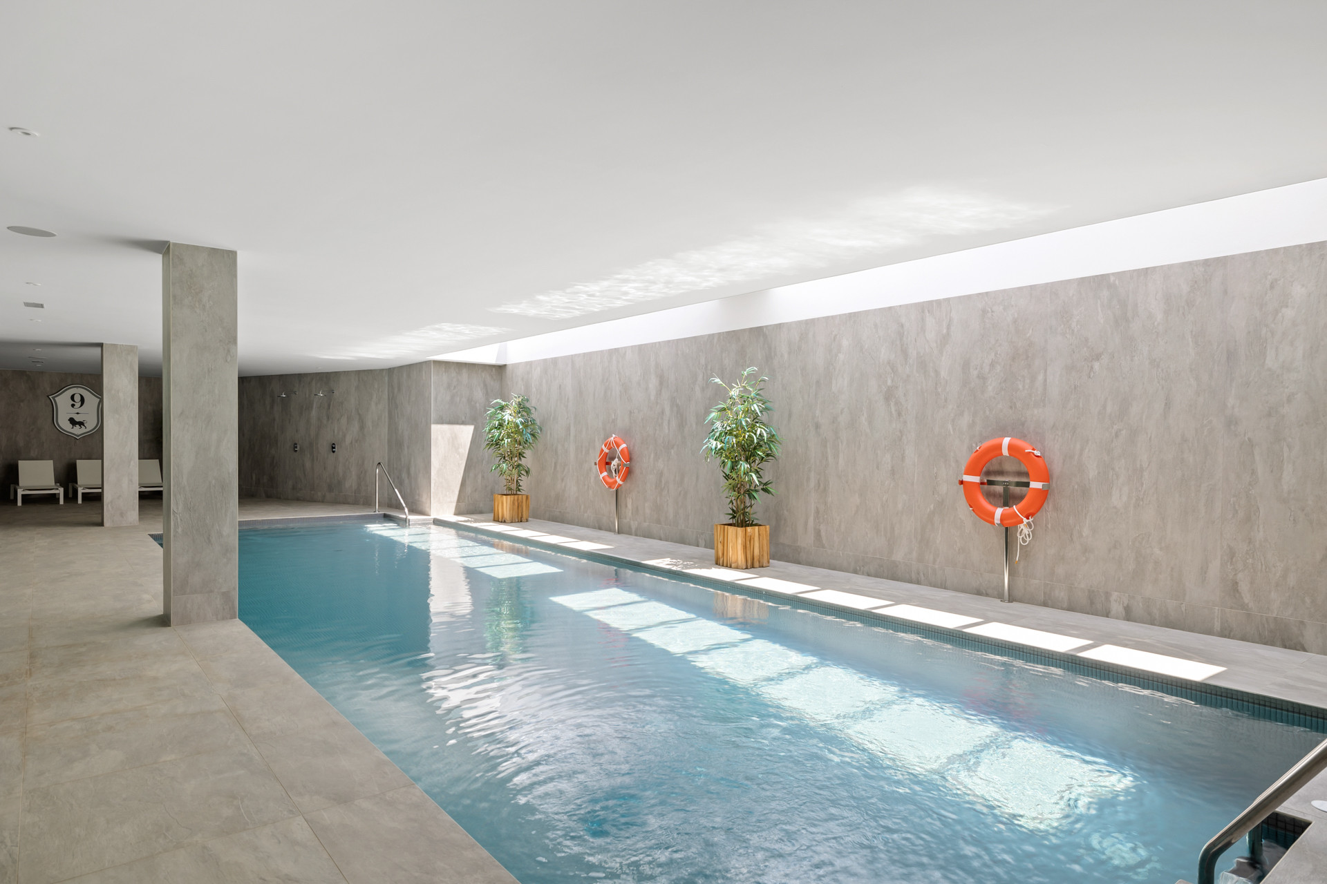 Indoor swimming pool with concrete walls, two orange life preservers, and plants in planters.