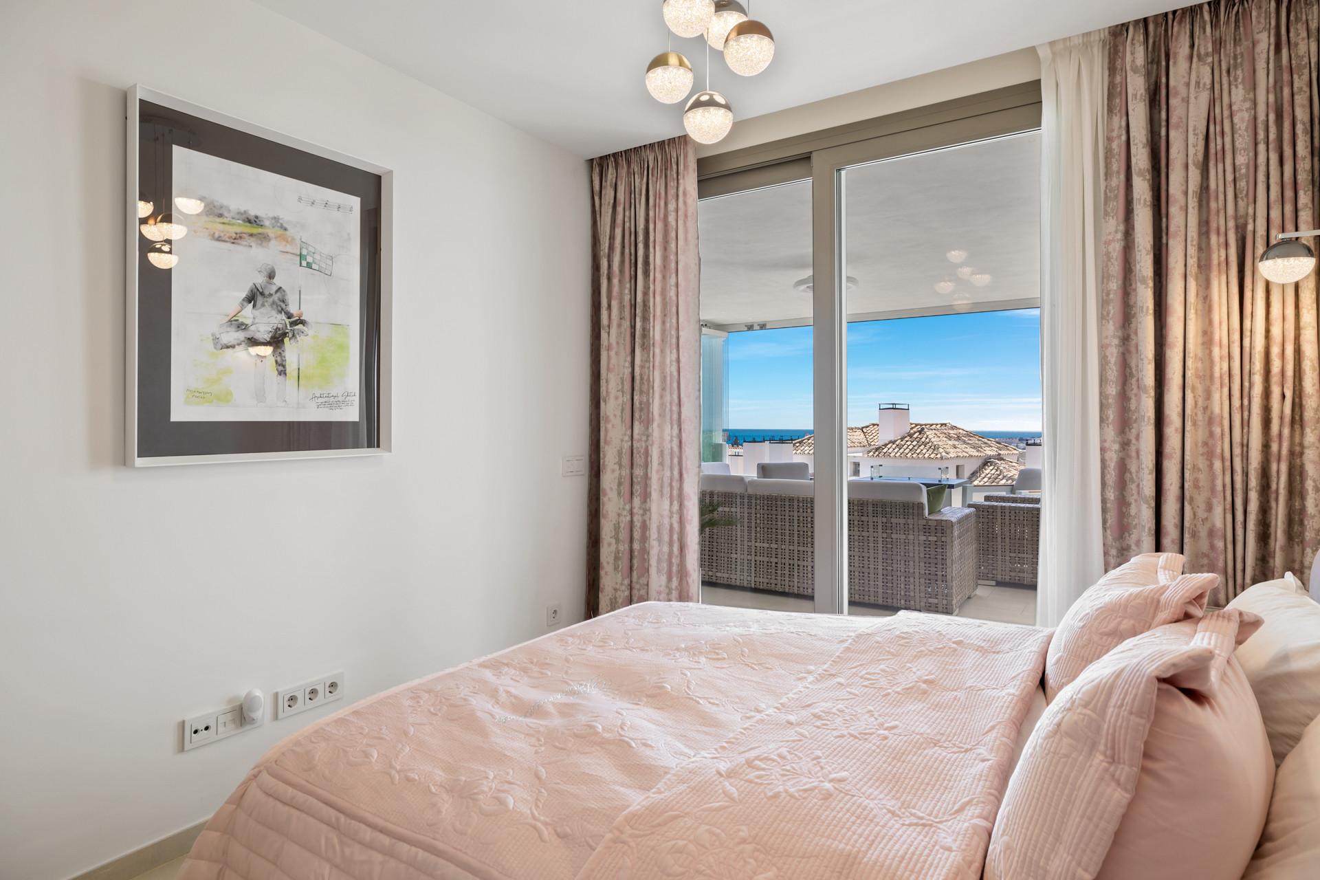 Bedroom with a large artwork, pink quilted bedspread, and a view of the sea from the balcony.