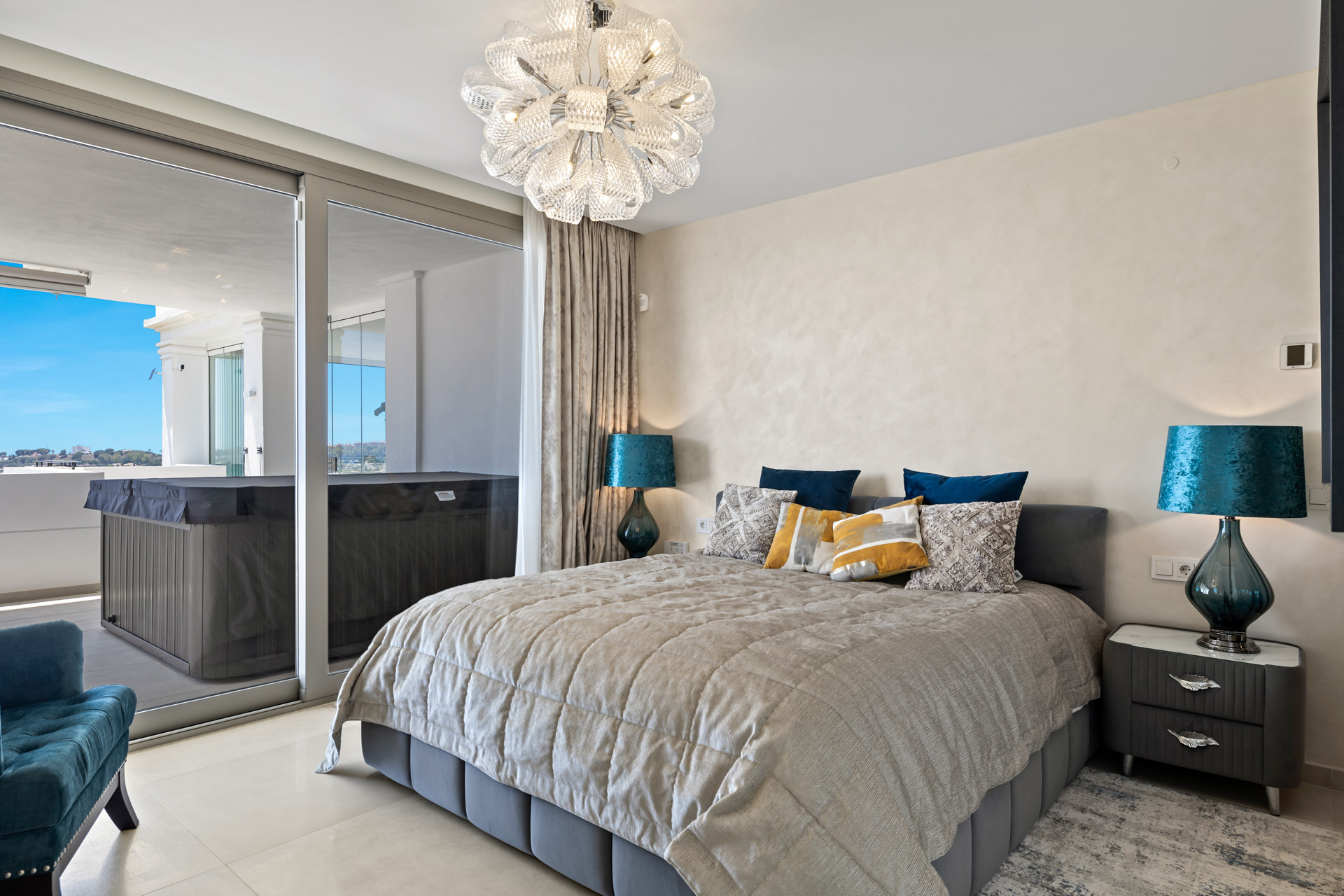 Bedroom with a large bed, two nightstands with blue lamps, and sliding doors to a balcony with a hot tub.